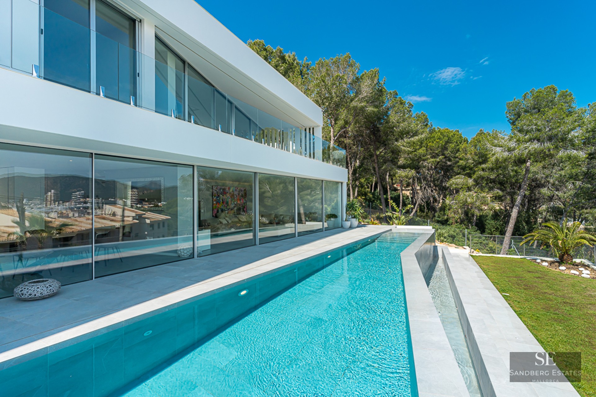 A sleek modern white villa with a long turquoise lap pool, floor-to-ceiling glass windows, and lush green trees.