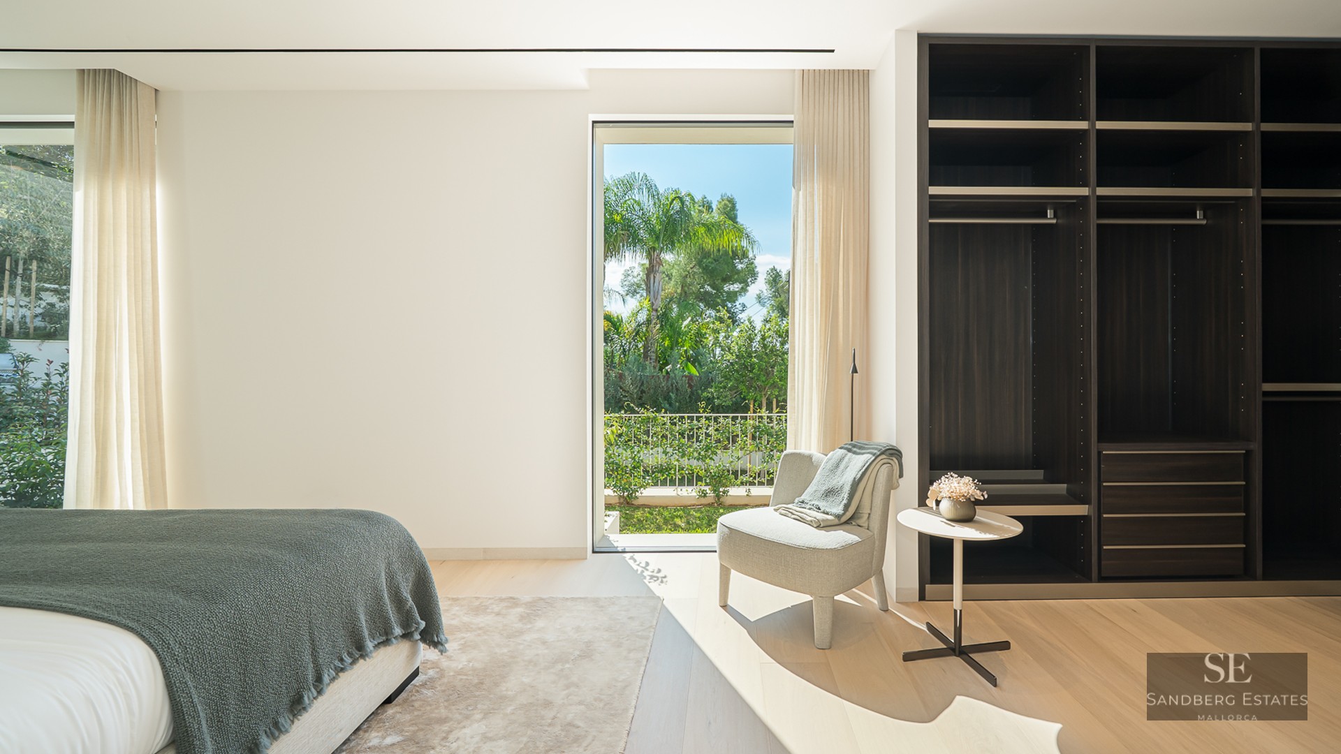 Bright modern bedroom featuring a large dark wood closet, a lounge chair, and a floor-to-ceiling view of a tropical garden.