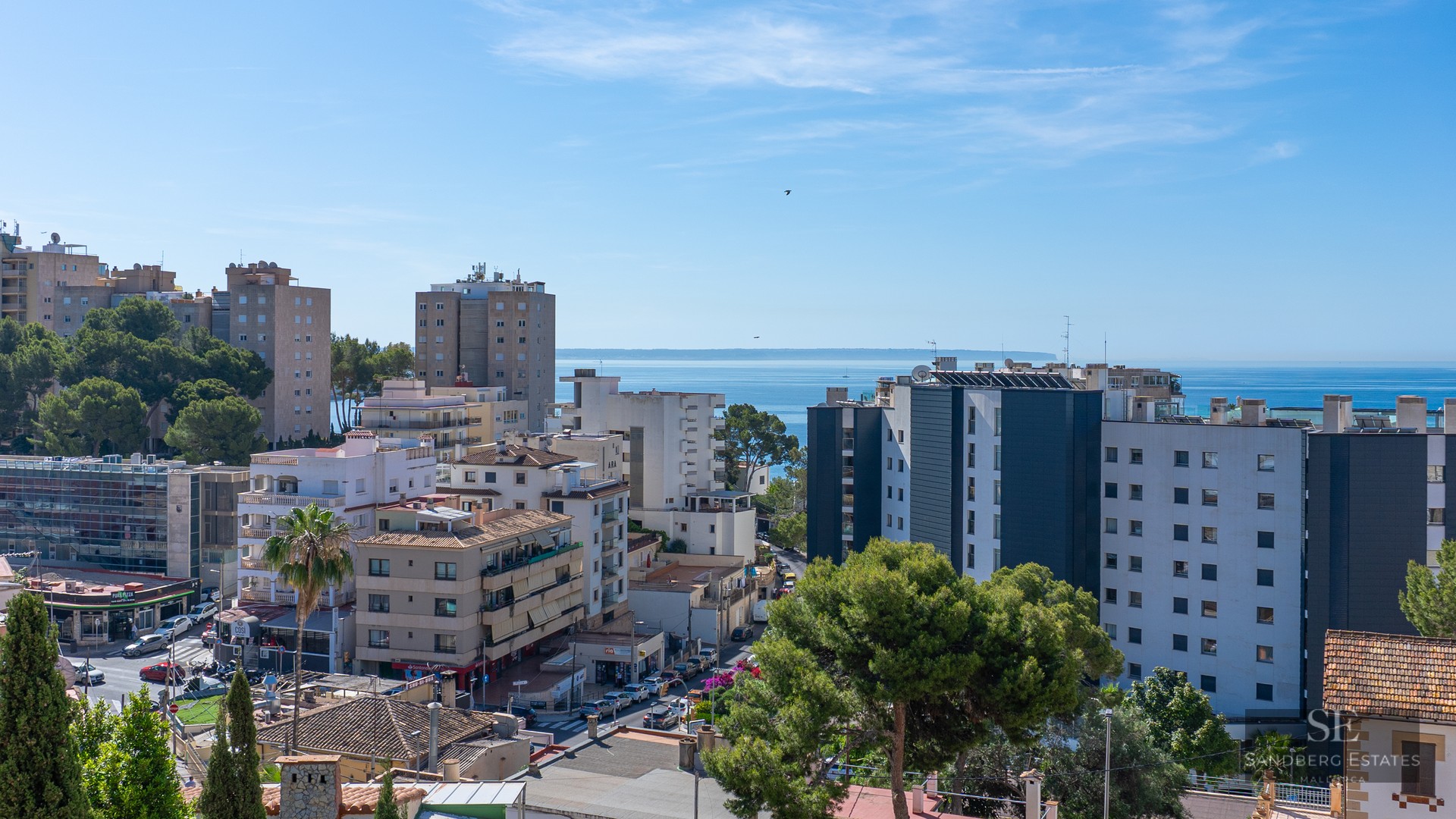 Elevated view of a Mediterranean coastal town with various buildings, green trees, and the blue sea on the horizon.