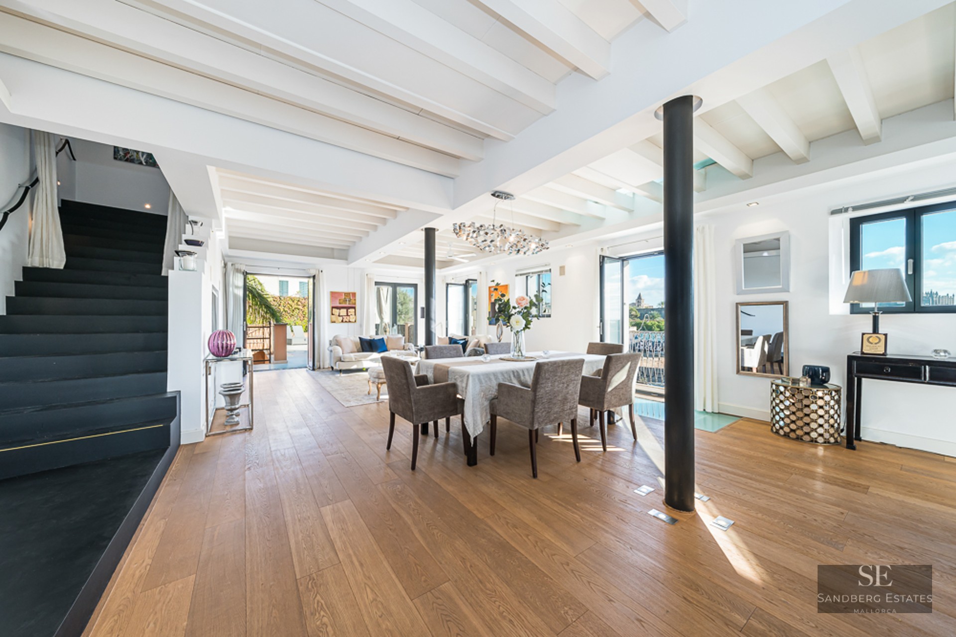 Bright open-plan room with wooden floors, white beamed ceiling, dining set, and a black staircase on the left.