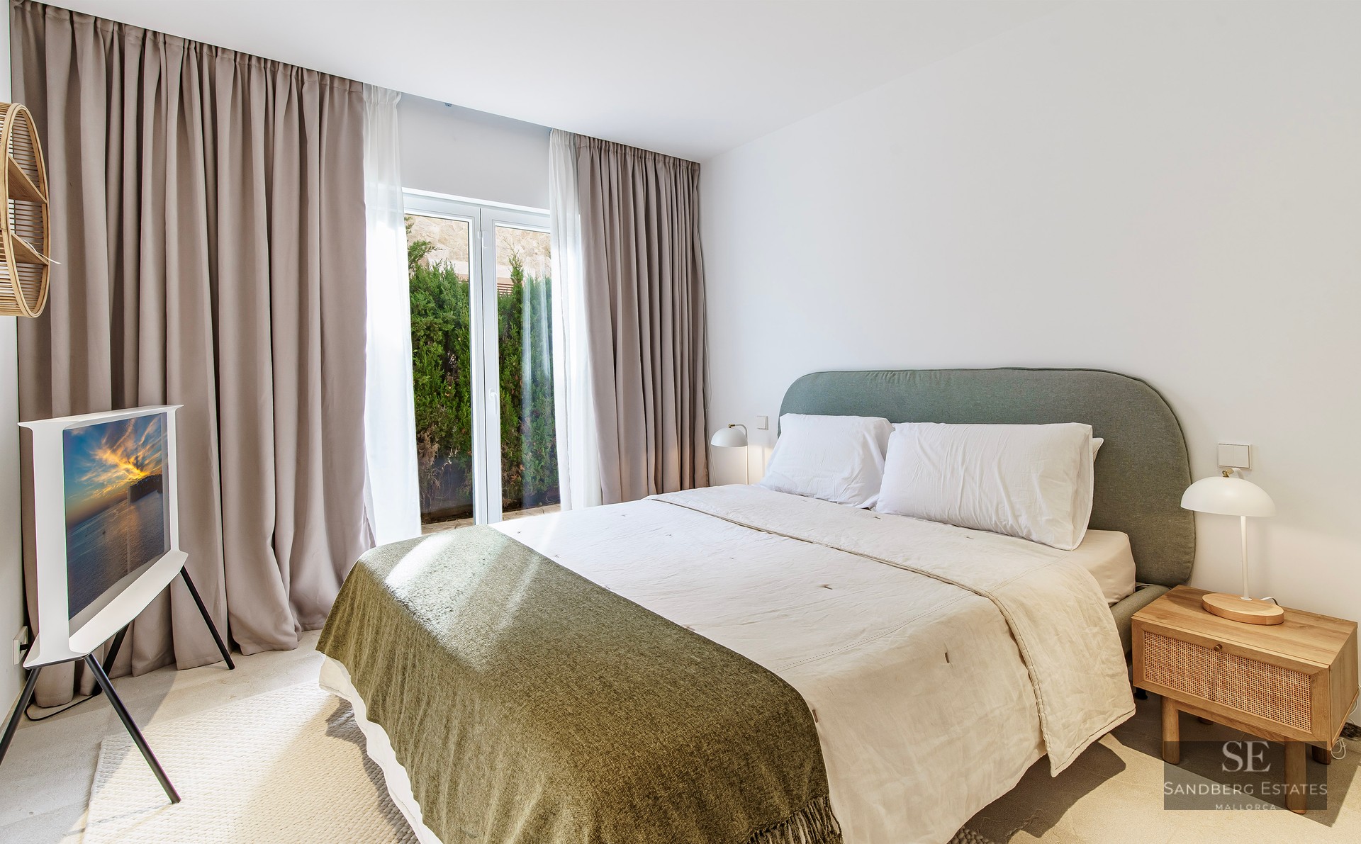 Bright bedroom with large bed, gray headboard, wooden nightstands, and floor-to-ceiling windows with beige curtains.