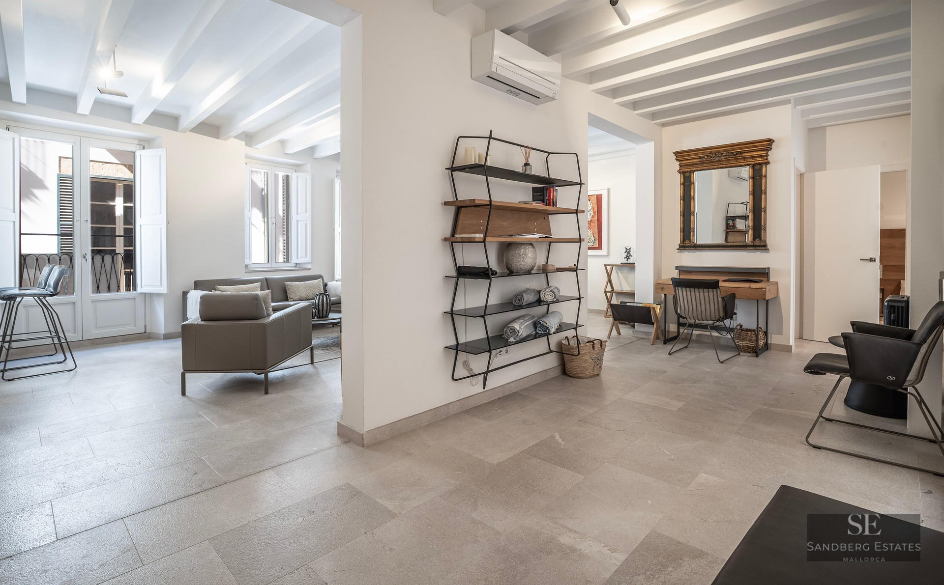 Modern interior with stone floors, white beams, grey sofa, and a study nook with a gold-framed mirror.