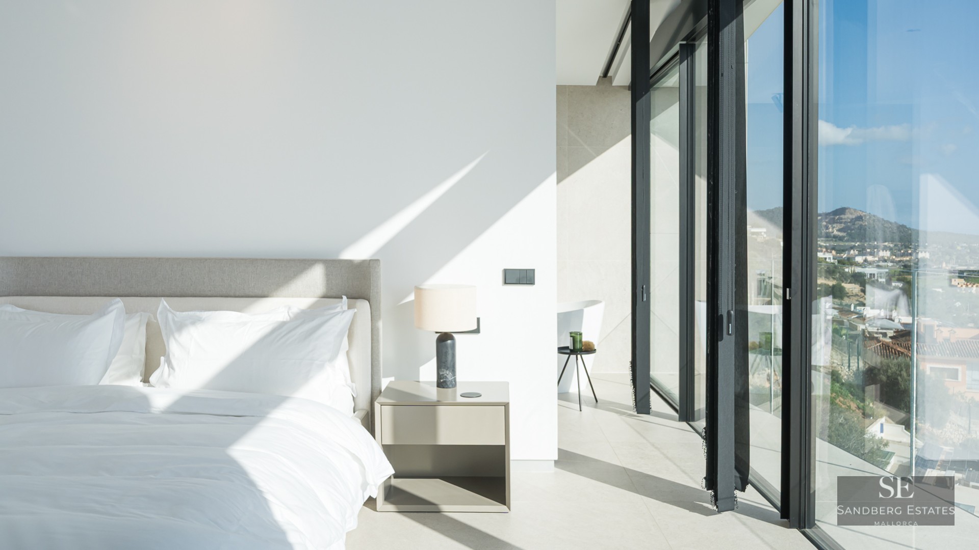 Bright minimalist master bedroom with floor-to-ceiling glass windows and white linens under natural sunlight.