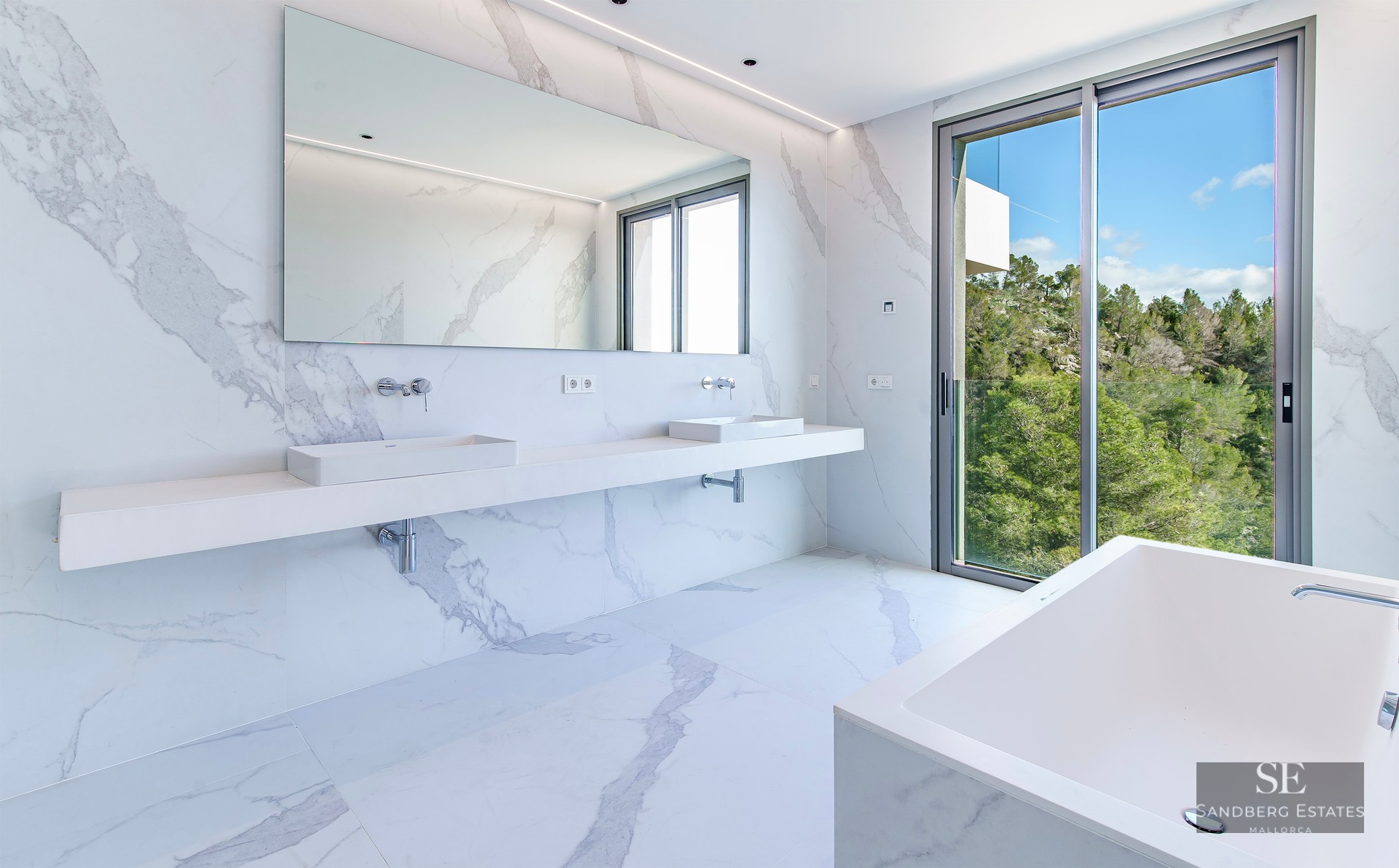 Bright master bathroom with white marble walls and floors, double vanity, bathtub, and large window view of forest.