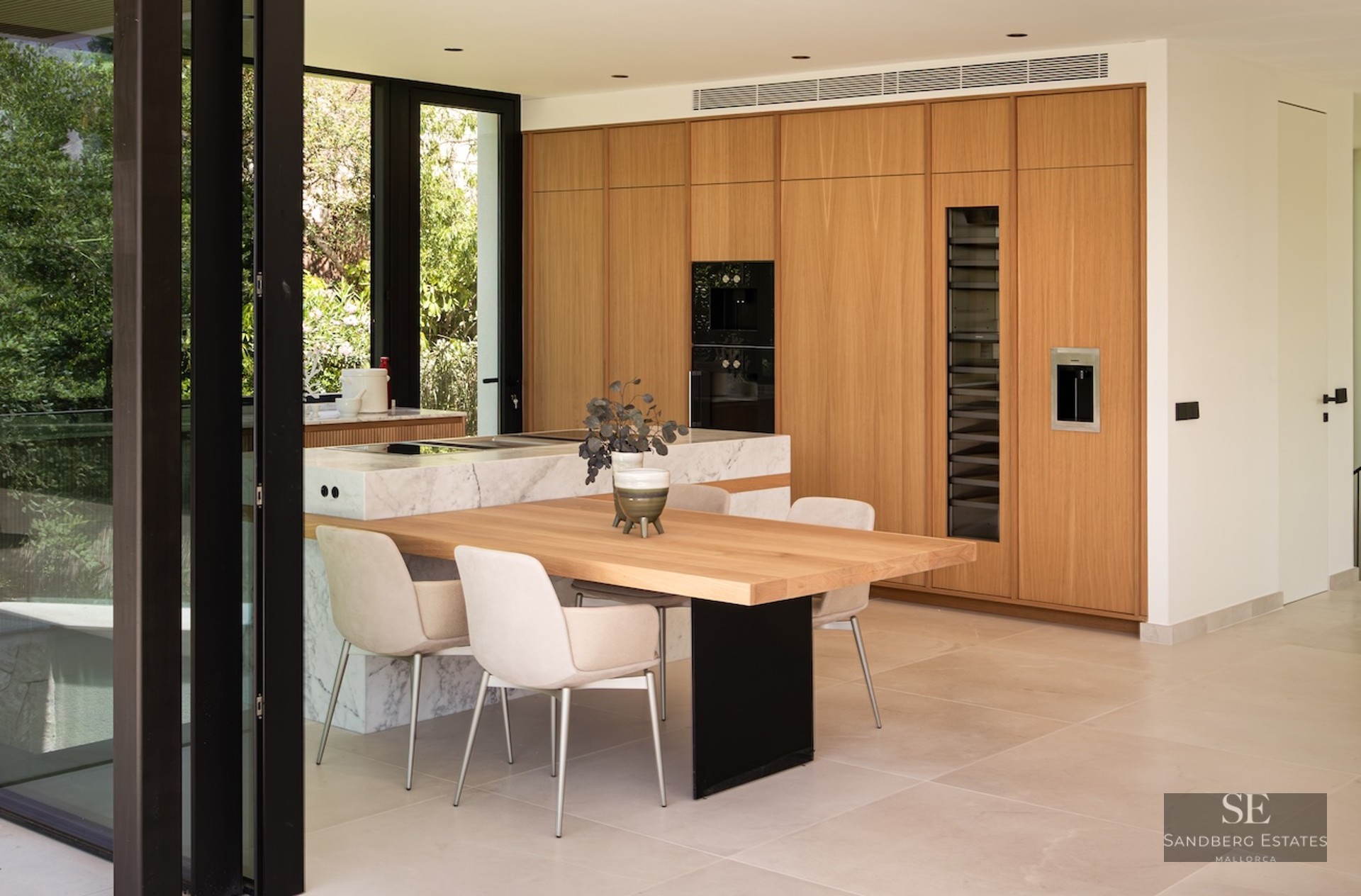 Modern kitchen with wood cabinets, marble island, integrated dining table, and large glass windows showing garden views.