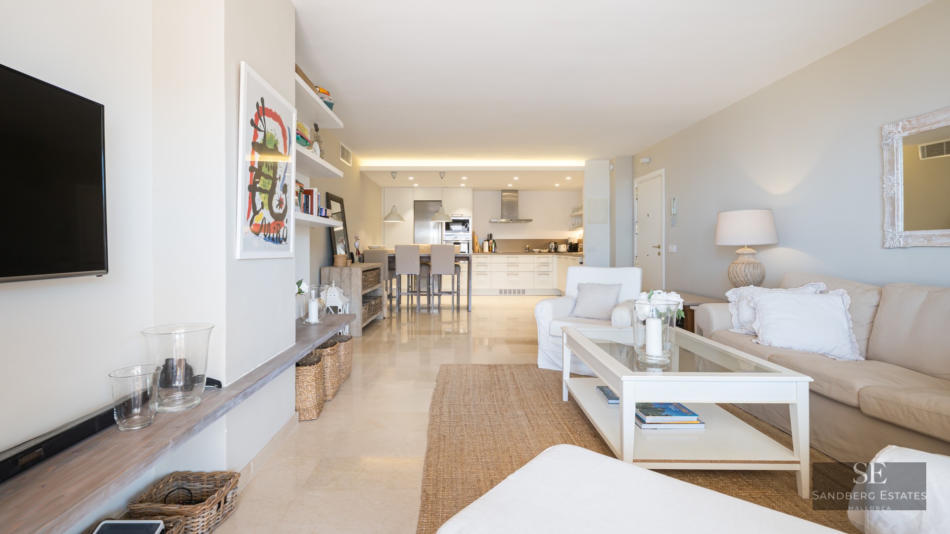 Bright living room with cream sofa, white coffee table, and open kitchen in the background with a breakfast bar.
