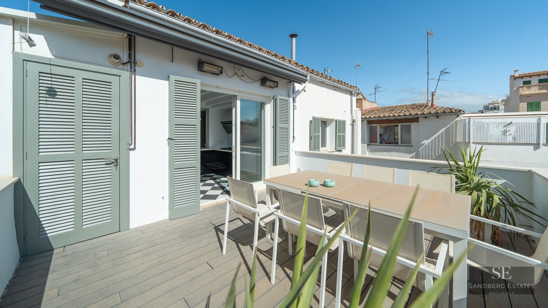 Sunny terrace with a dining table for six, sage green shutters, and wooden decking under a clear blue sky.