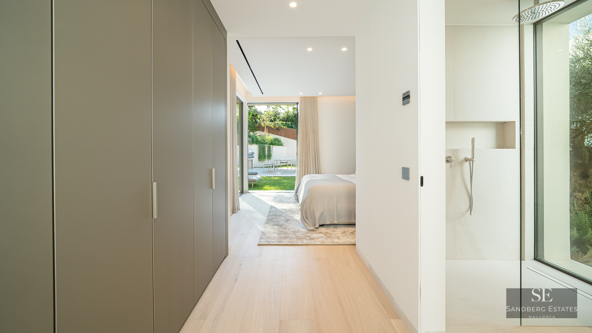 Hallway with built-in closets leading to a modern bedroom with a glass shower and view of a sunny garden.