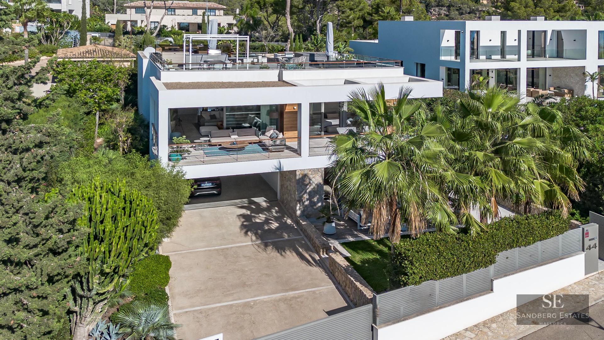 Vue architecturale d'une villa moderne blanche avec terrasses, palmiers et murs en pierre sous une lumière vive.
