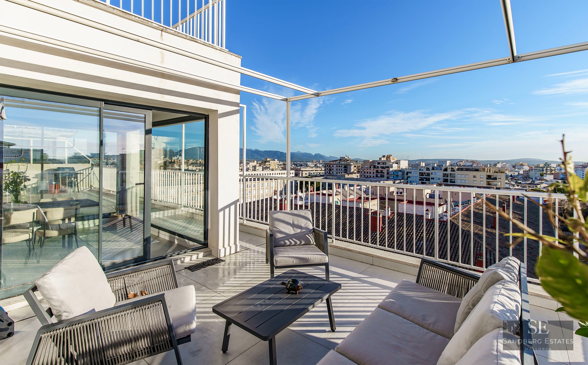 Spacious white terrace with modern outdoor lounge furniture overlooking a cityscape and distant mountains under a blue sky.