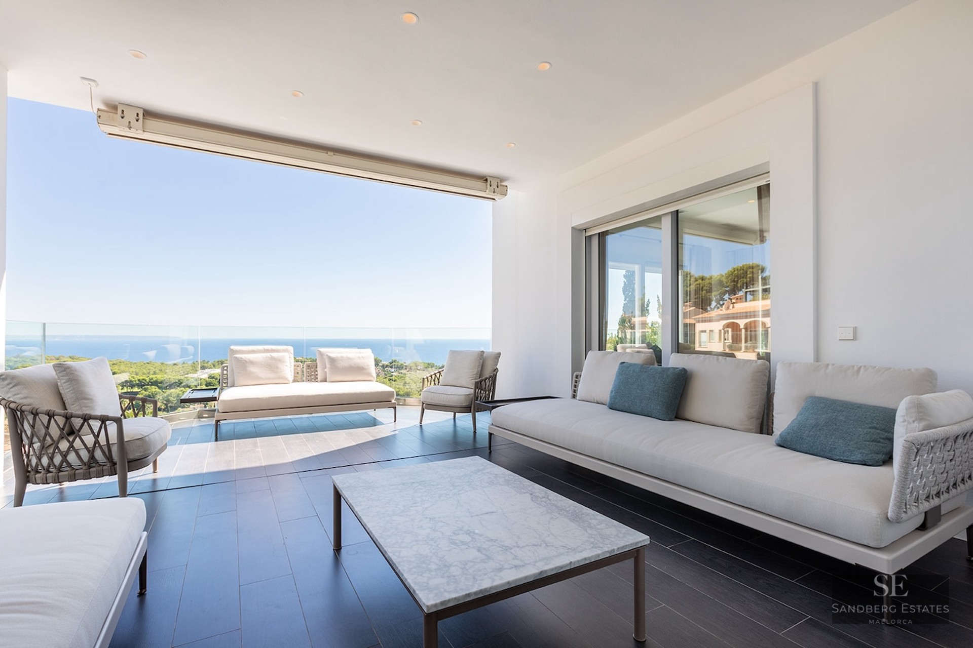 Modern covered terrace with white sofas, a marble coffee table, and a glass railing overlooking the Mediterranean sea.