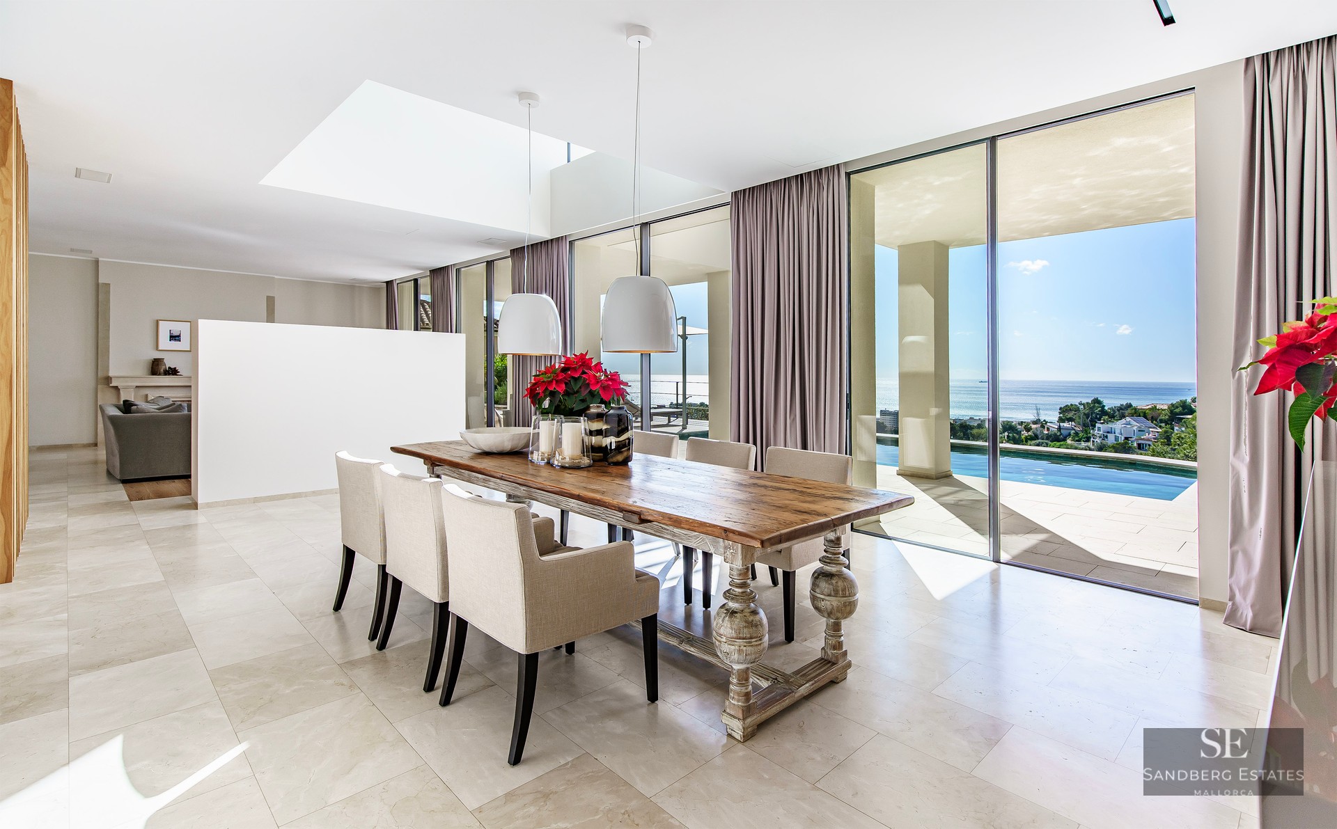 Spacious dining room with a large wooden table, cream chairs, and floor-to-ceiling windows overlooking a sea and pool.