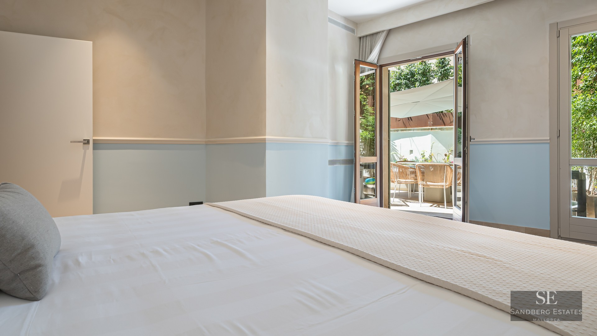 Bedroom with white linens and light blue walls looking out onto a sunny terrace through open French doors.