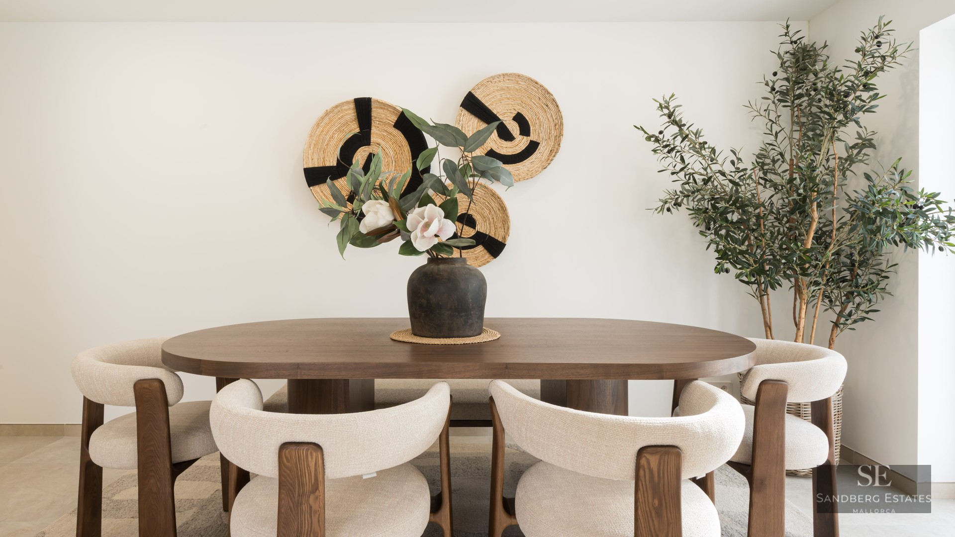 Dining room featuring an oval wooden table, cream chairs, woven wall art, and a large indoor olive tree.