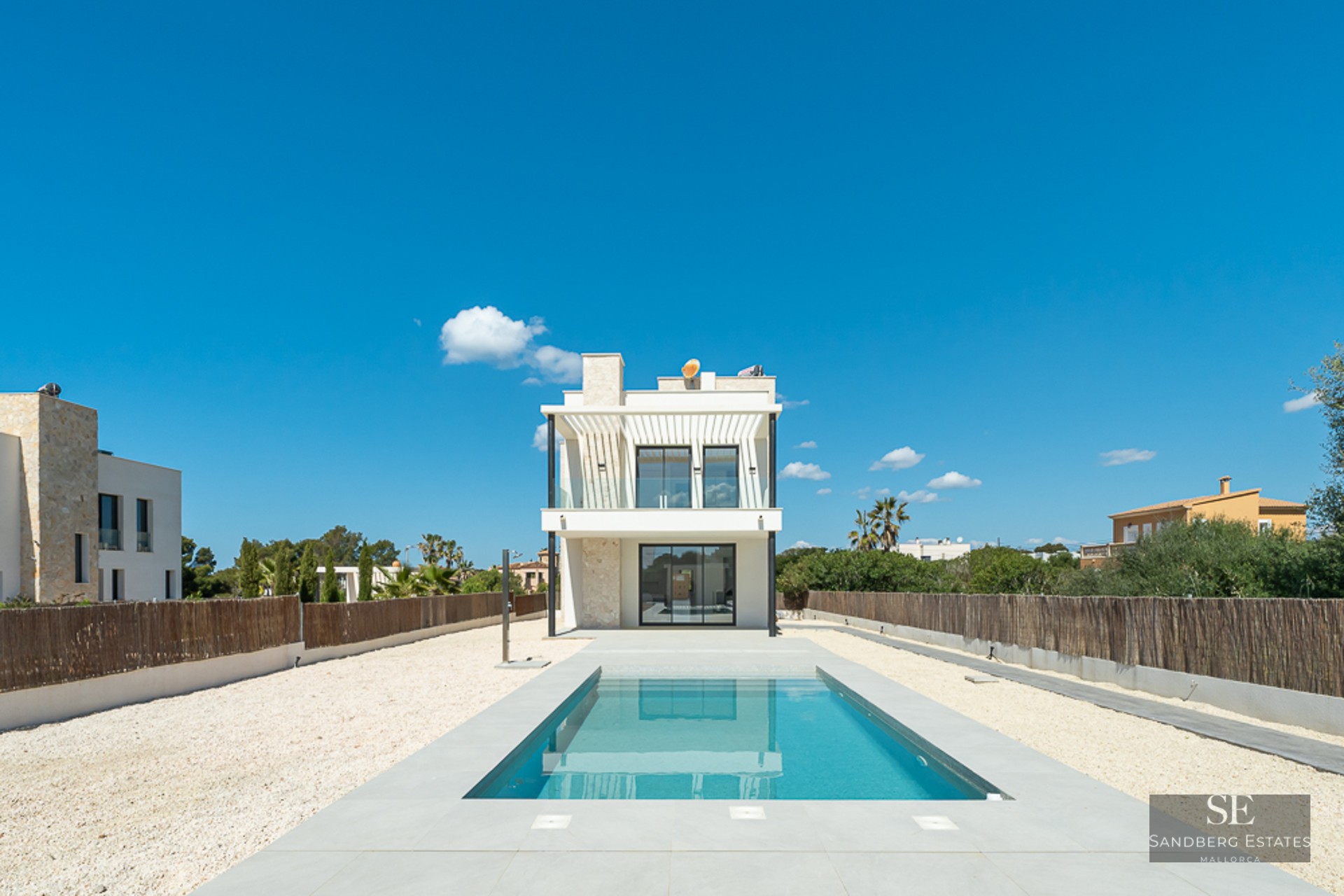 A rectangular blue swimming pool leading to a modern white two-story villa under a clear blue sky.