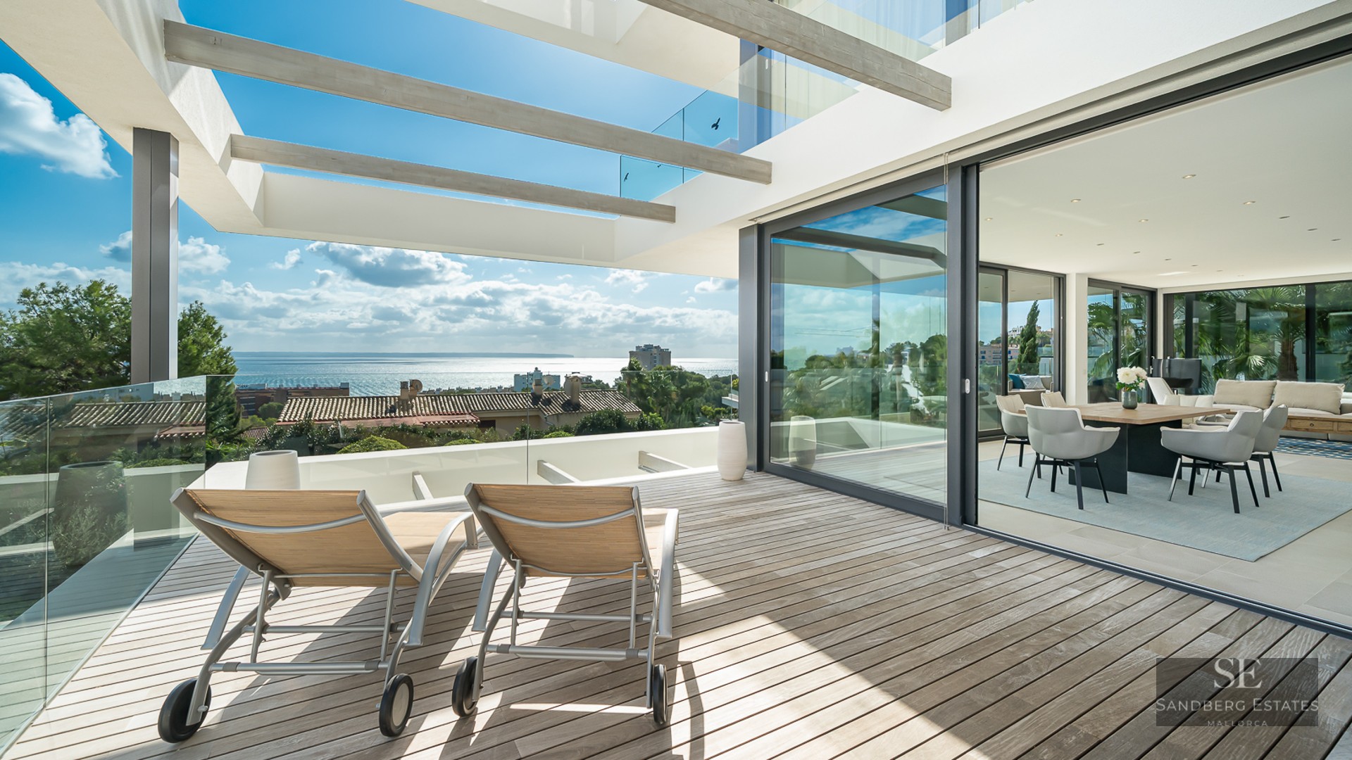 Sun loungers on a wooden terrace with glass railings overlooking the sea, adjacent to an open-plan dining room.