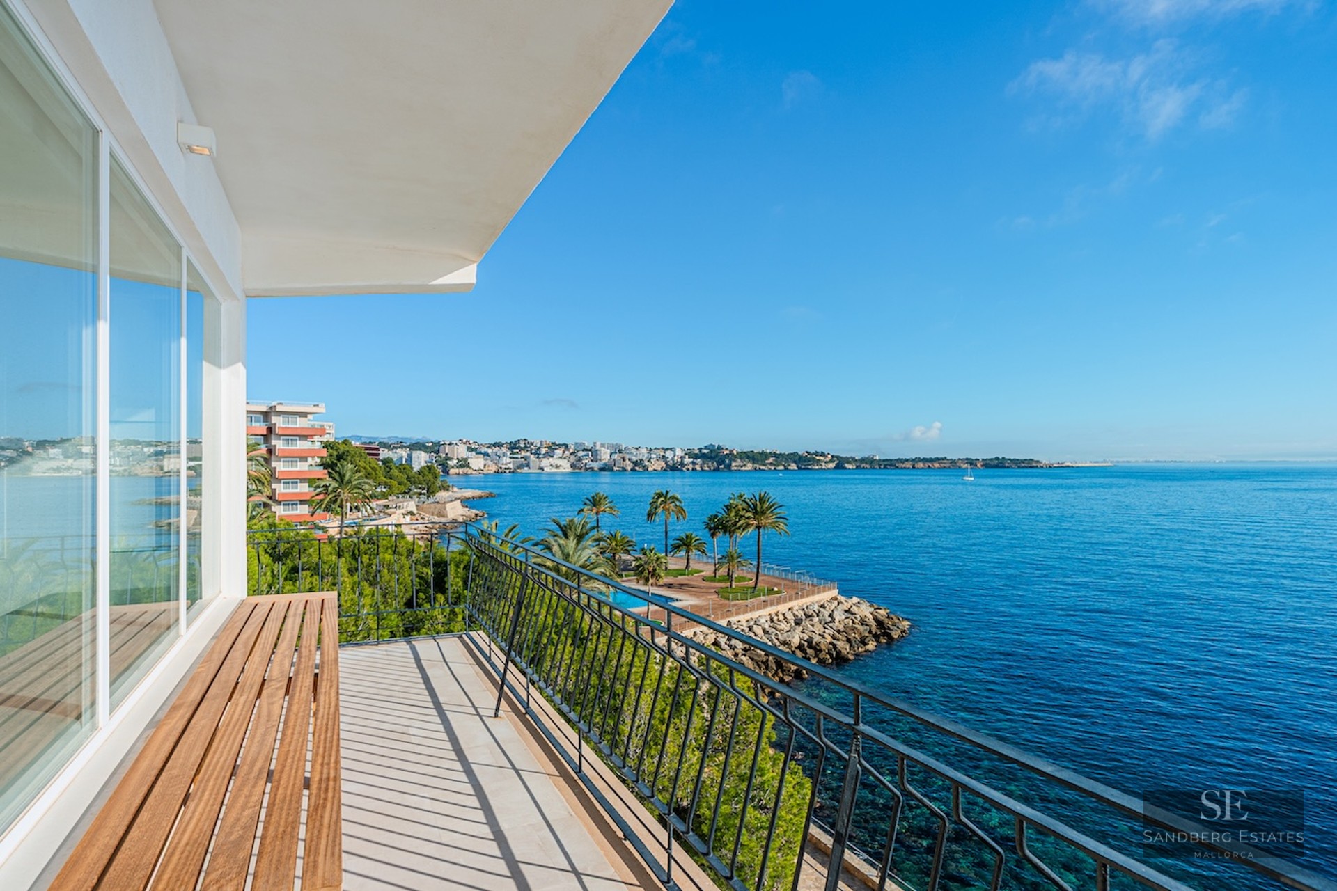 Balcony with wooden bench and black railing offering a panoramic view of the blue Mediterranean sea and coastline.