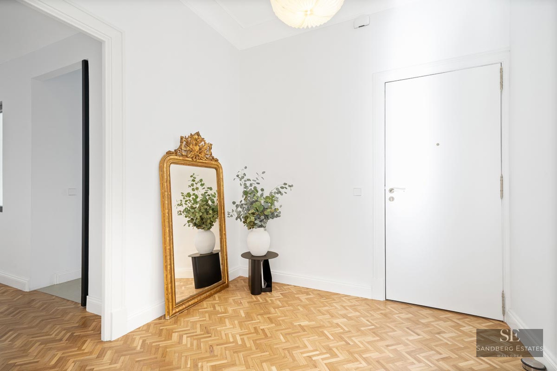 Bright foyer with white walls, herringbone wood floors, a large gold mirror, and a white door.