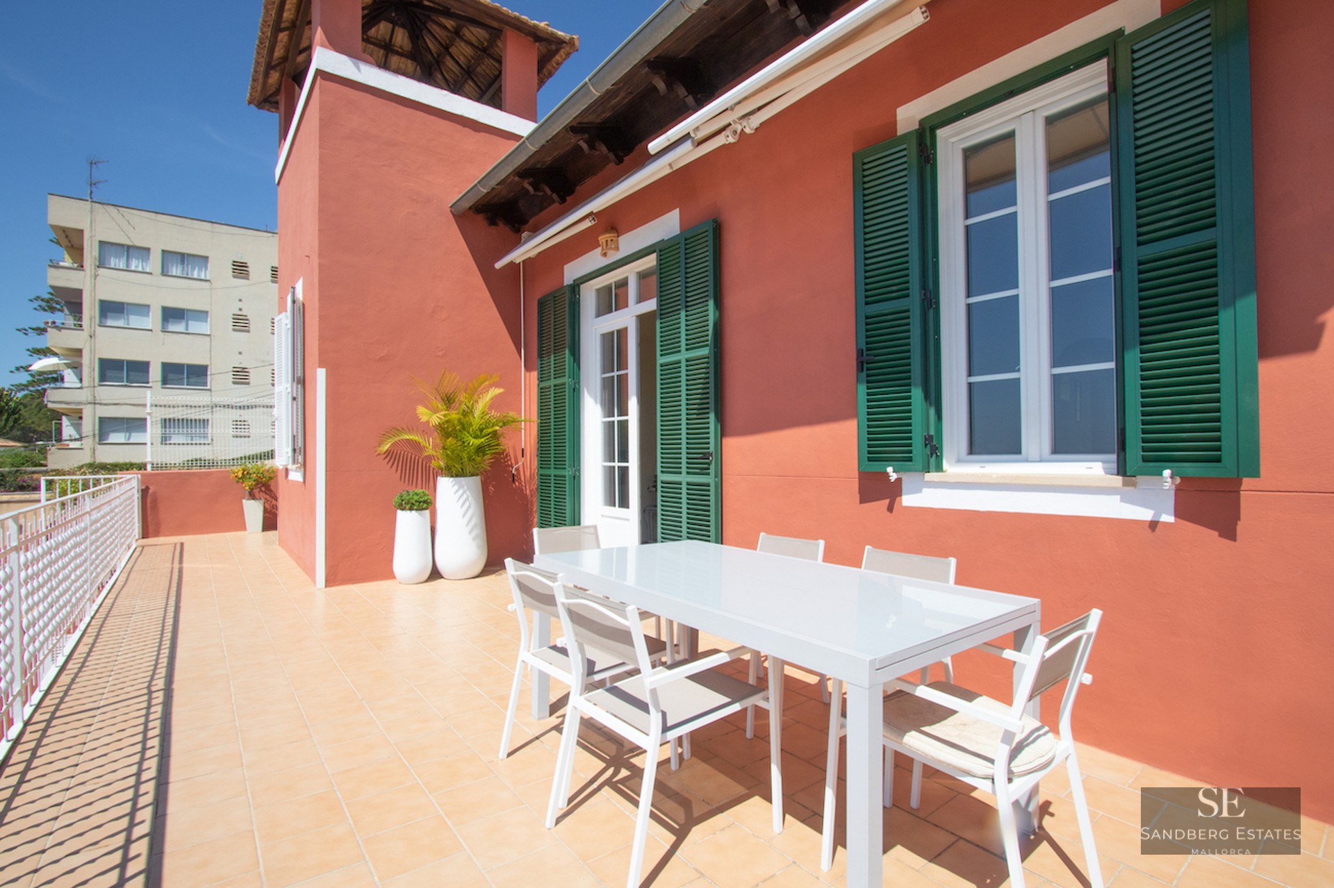 A sunlit terrace with terracotta walls, green shutters, and a white modern dining set under a clear blue sky.