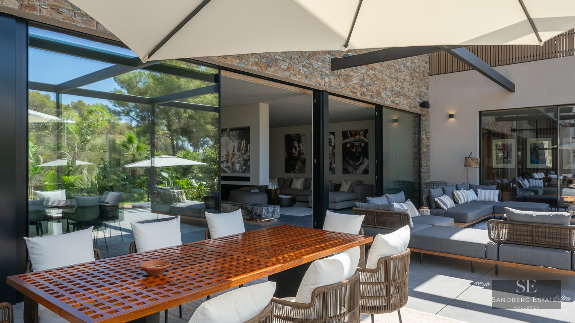 Modern terrace featuring a large wooden dining table, grey sofas, and floor-to-ceiling glass doors to the living room.