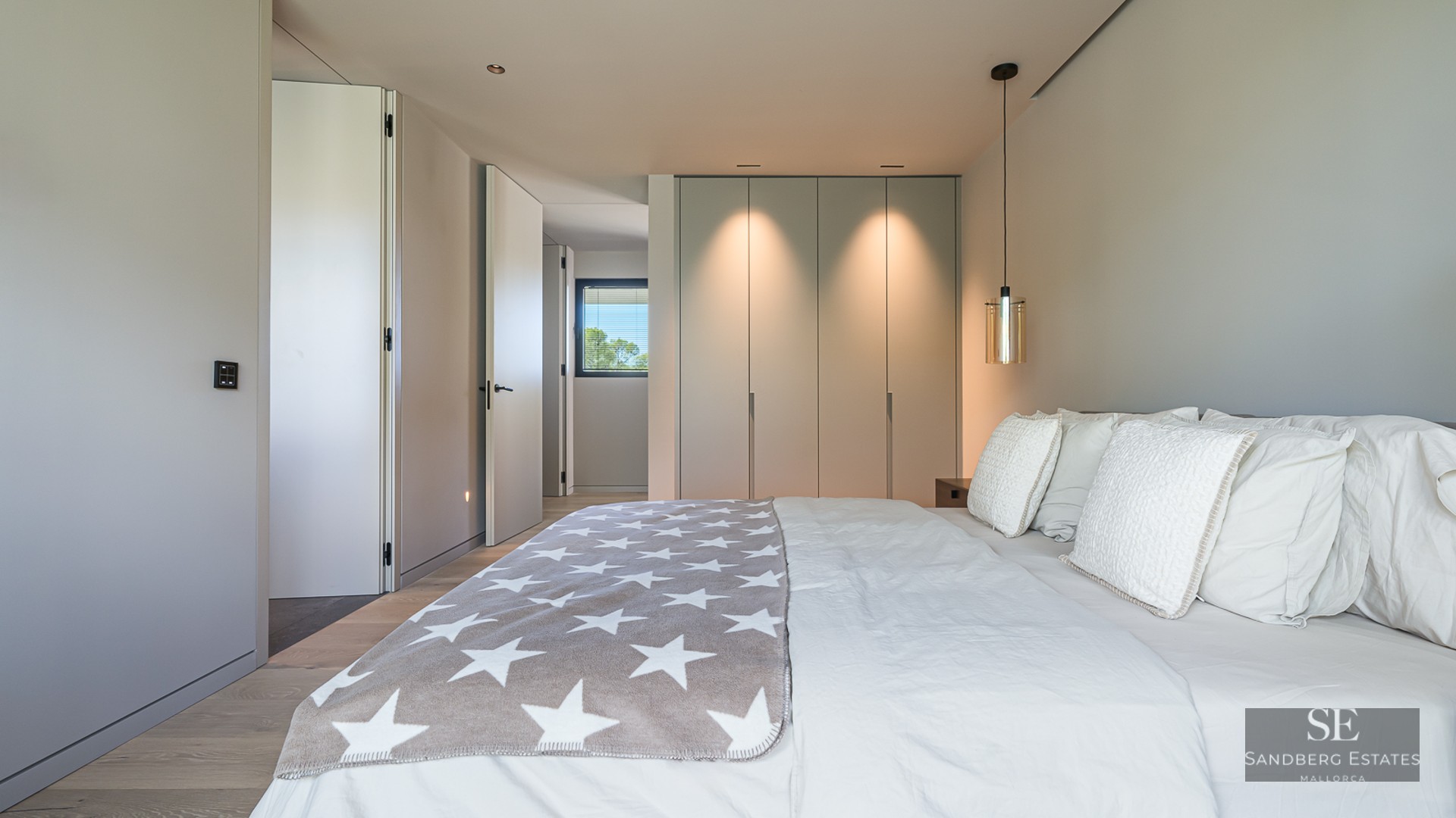 Bright modern bedroom featuring a large bed with white linens, grey star throw, and built-in closets with warm lighting.