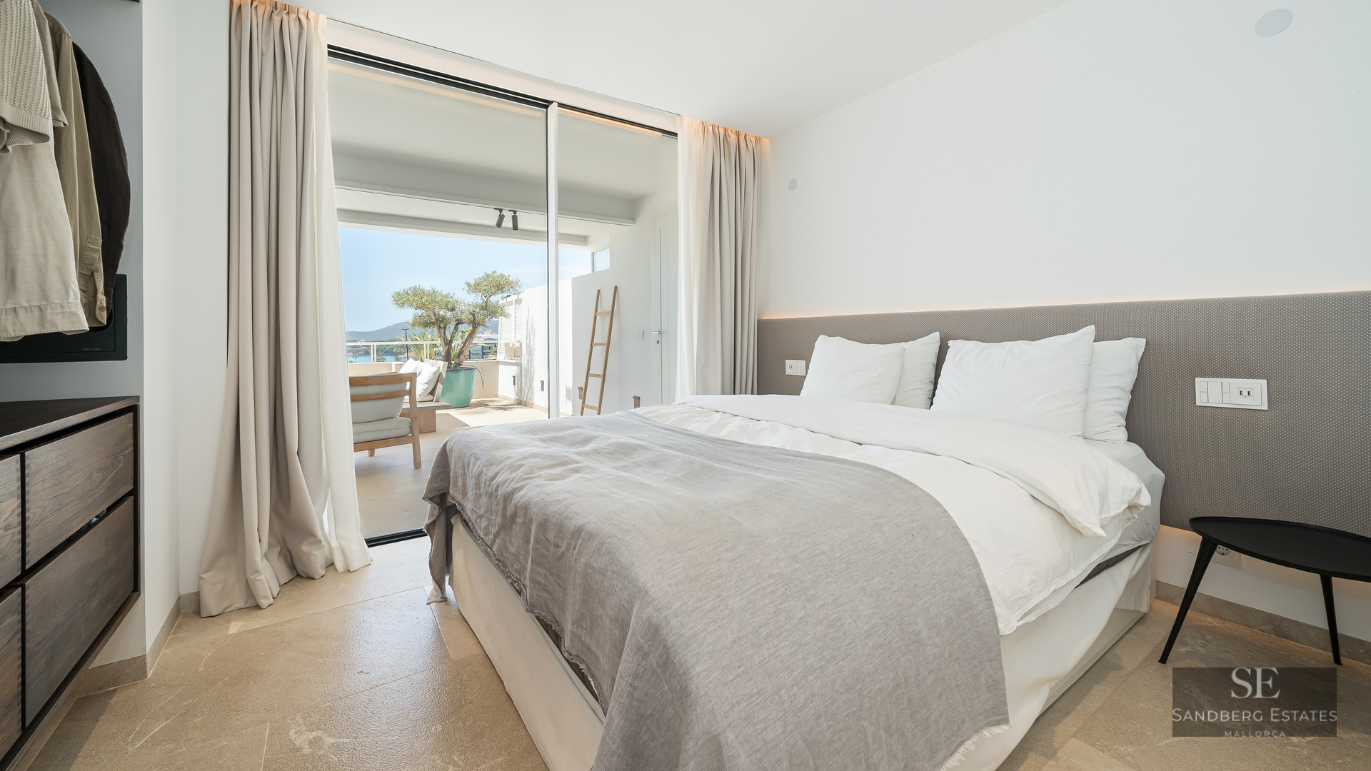 Bright modern bedroom with a large bed, stone floors, and floor-to-ceiling glass doors opening to a sunny terrace.
