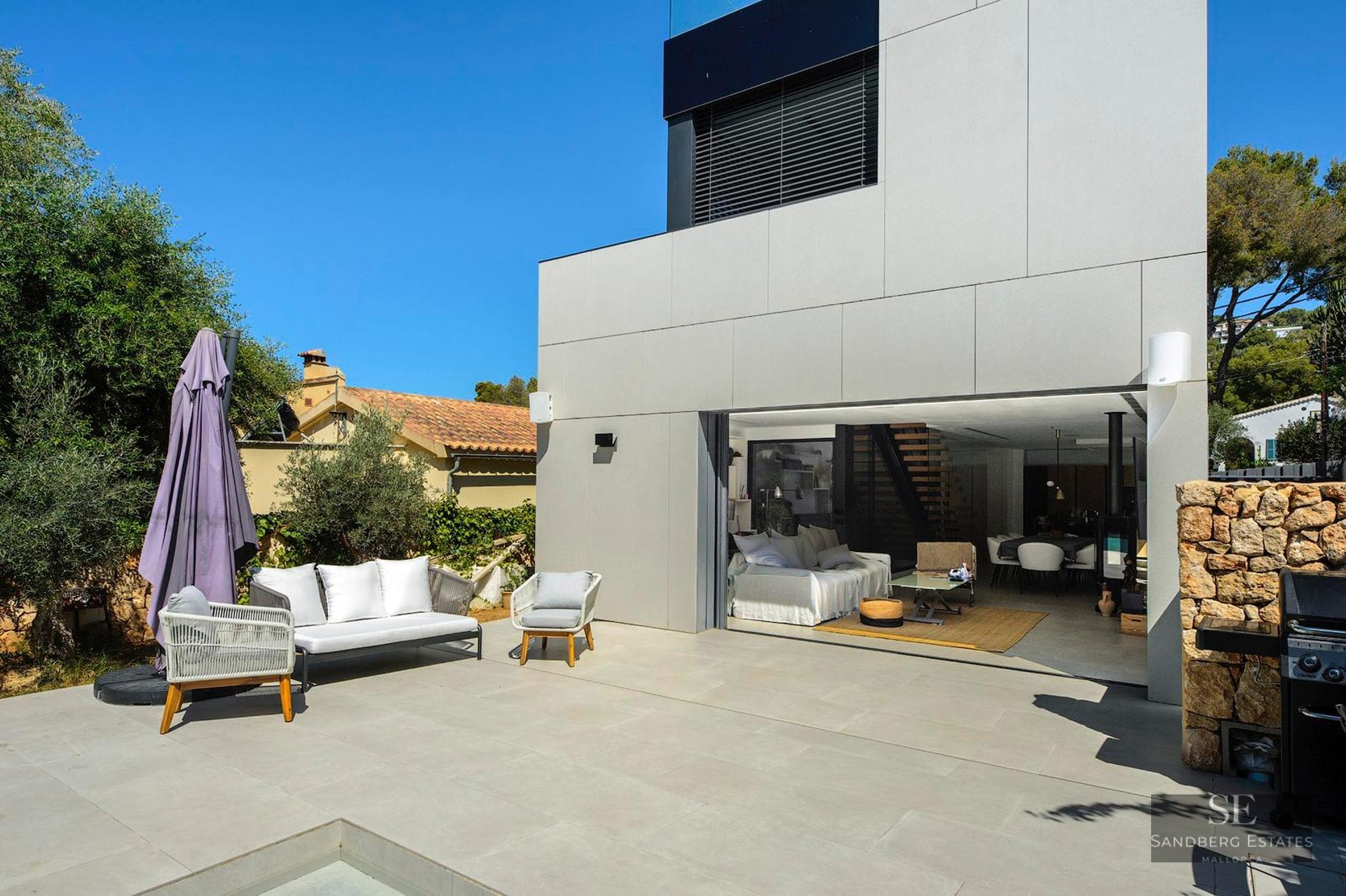 Modern outdoor terrace with white furniture next to a villa's open-plan living area and a stone wall.