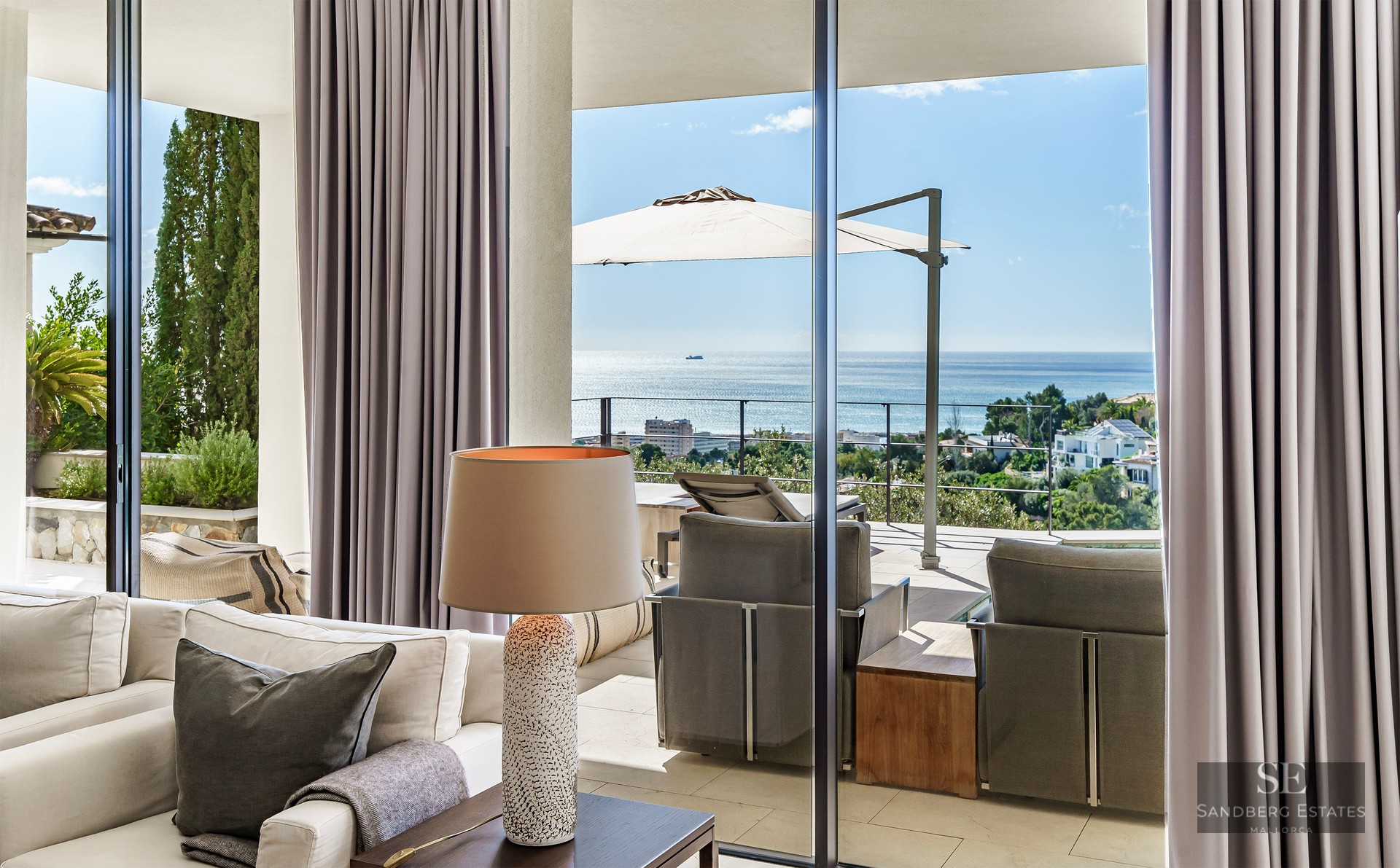 Elegant living room with beige sofas and floor-to-ceiling glass doors opening to a sunlit terrace with blue sea views.