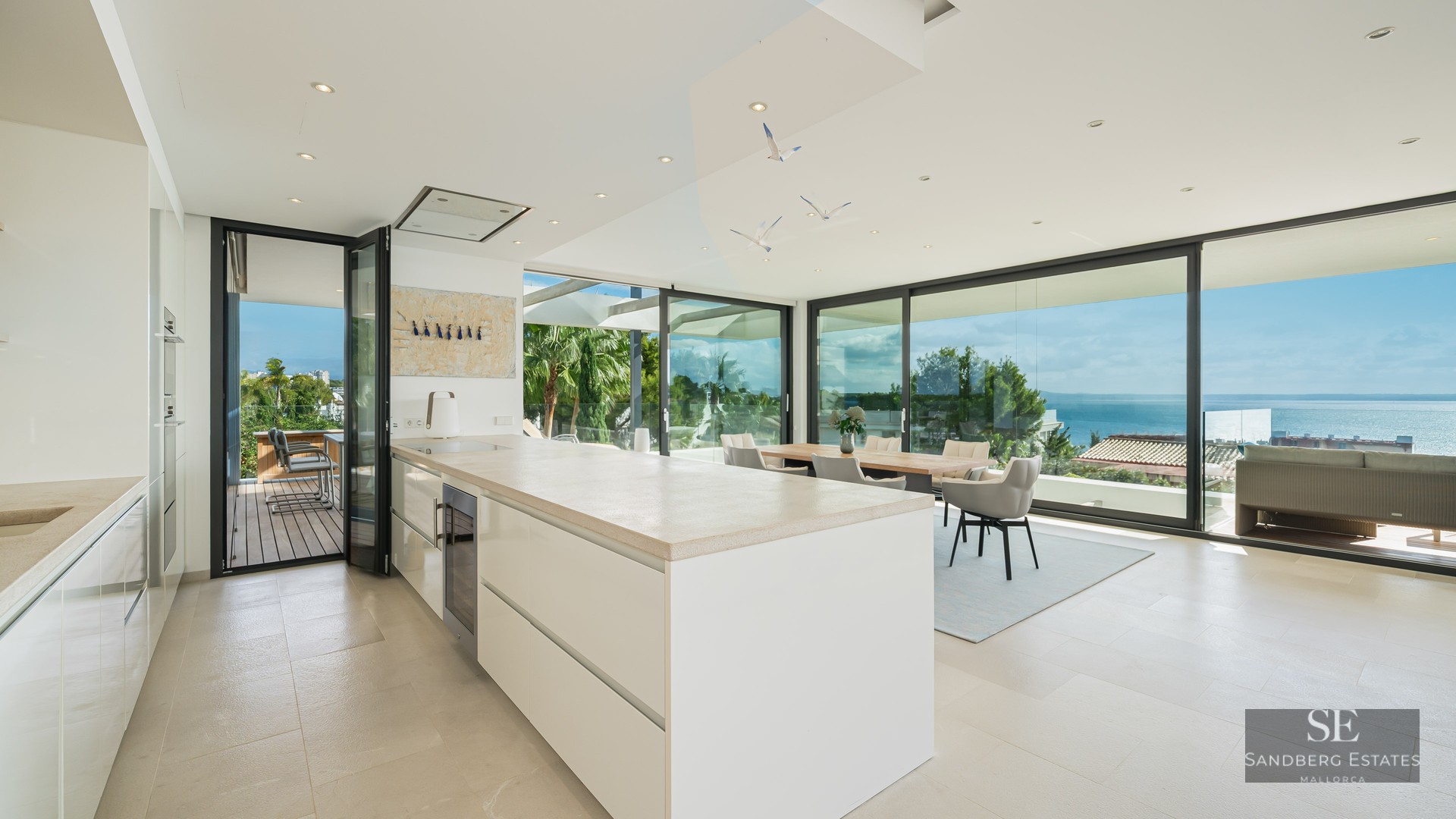 Bright modern white kitchen with large island, dining area, and floor-to-ceiling glass walls overlooking the ocean.