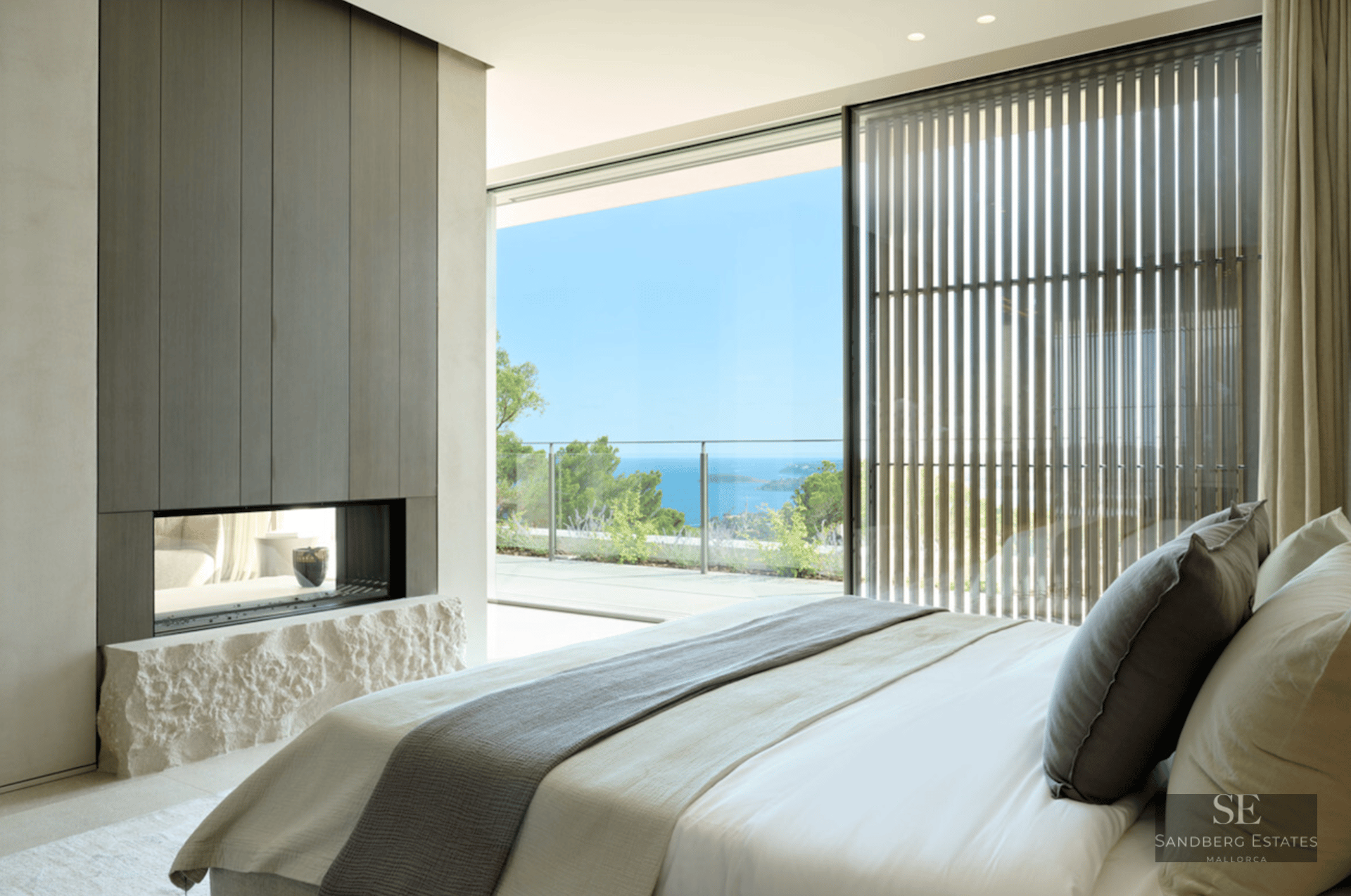 Luxury bedroom featuring floor-to-ceiling windows overlooking the ocean and a modern fireplace with a rough stone base.