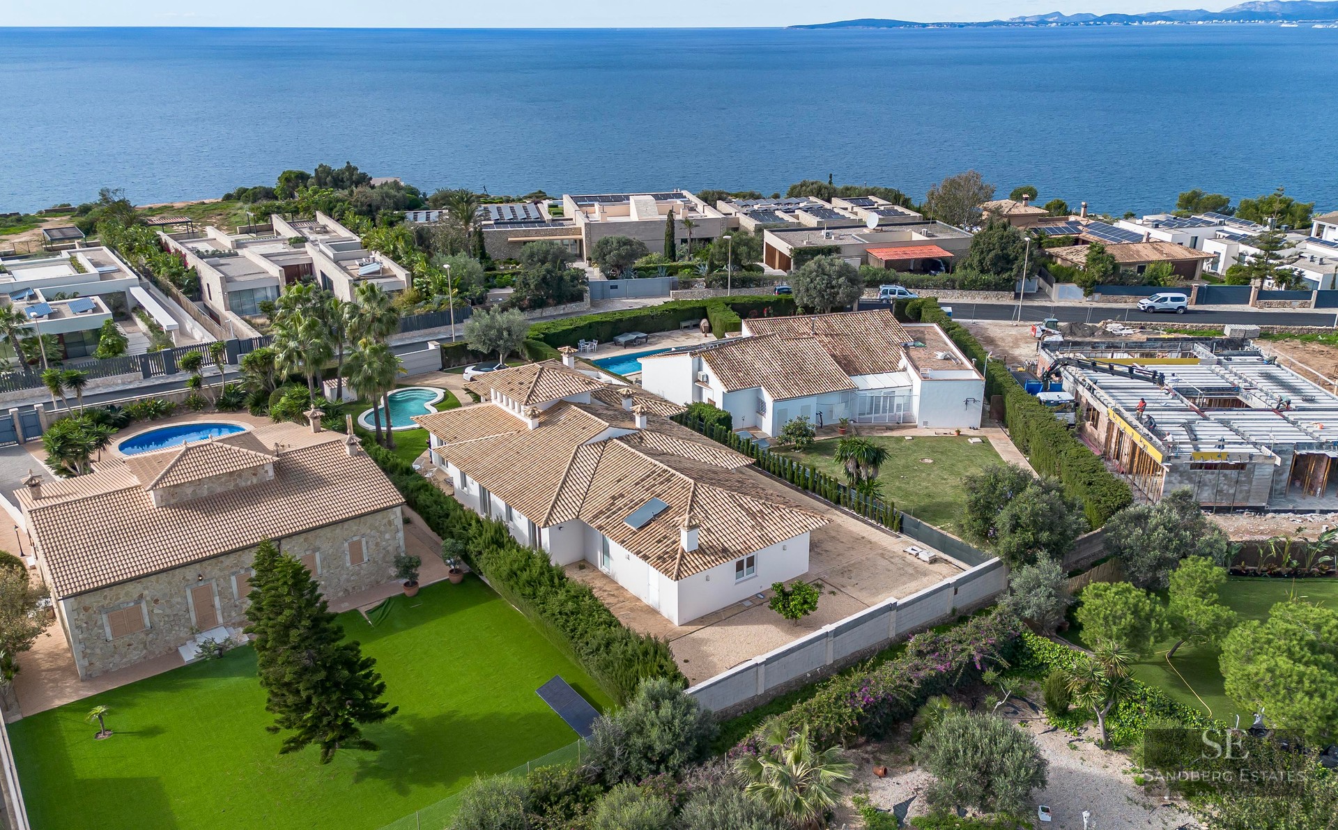 High-angle drone shot of a white villa with terracotta roof, green lawn, and the blue ocean in the background.