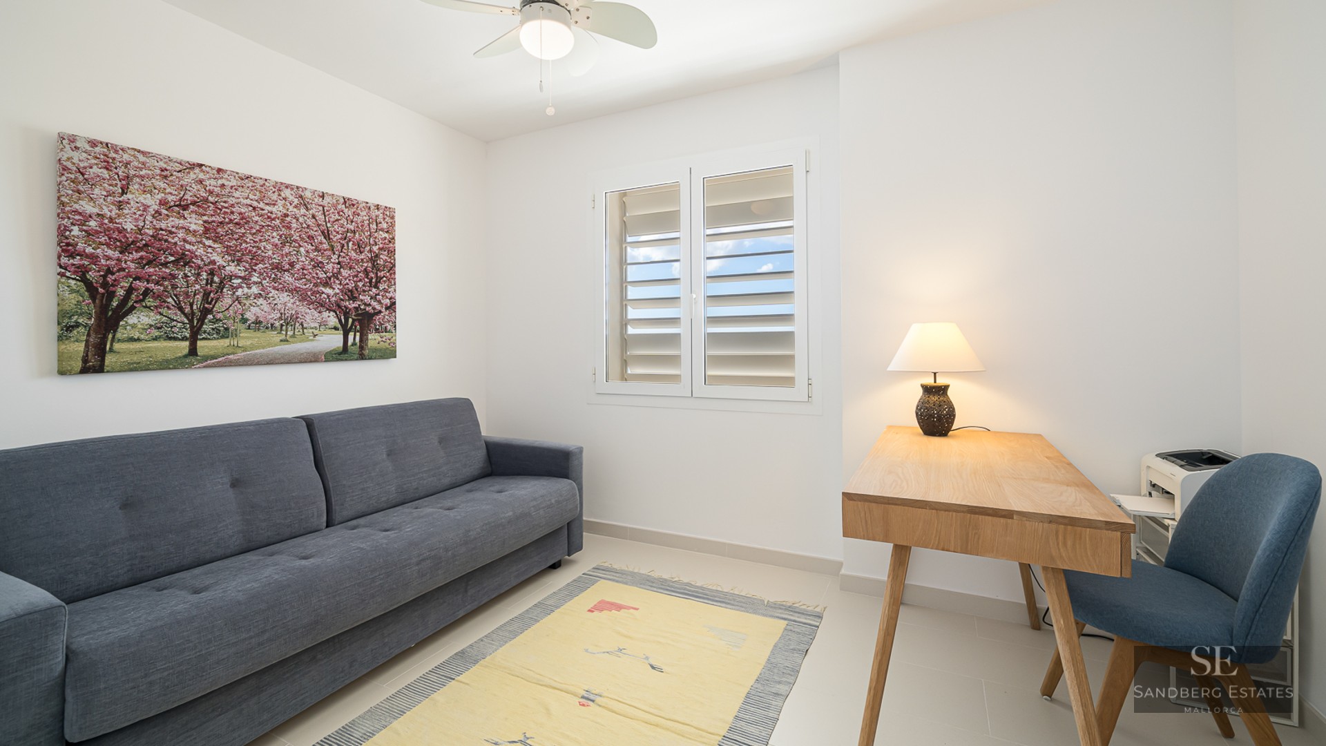 A minimalist room with a grey sofa, wooden desk with a lamp, a blue chair, and a window with white shutters.