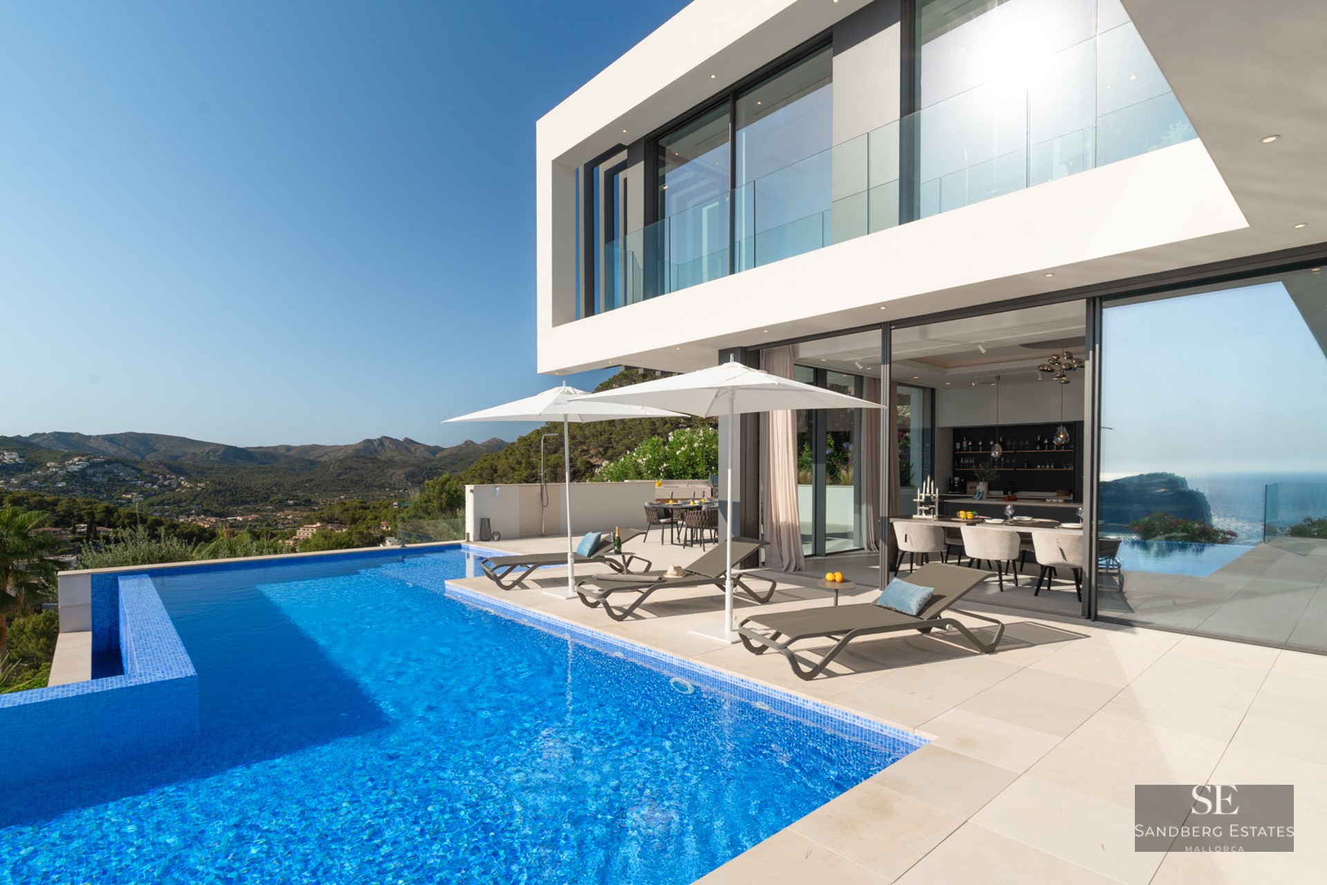 Luxury infinity pool and white modern villa with mountain views and designer loungers under white umbrellas.