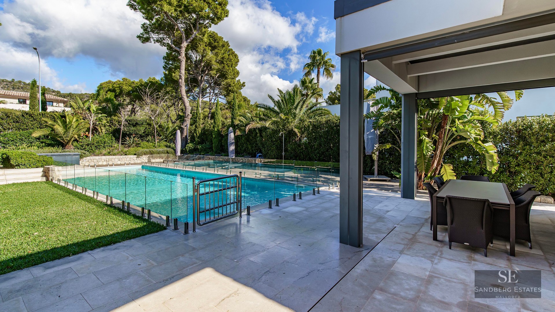 Piscine turquoise avec clôture en verre à côté d'une terrasse en pierre ombragée avec table et jardin tropical.