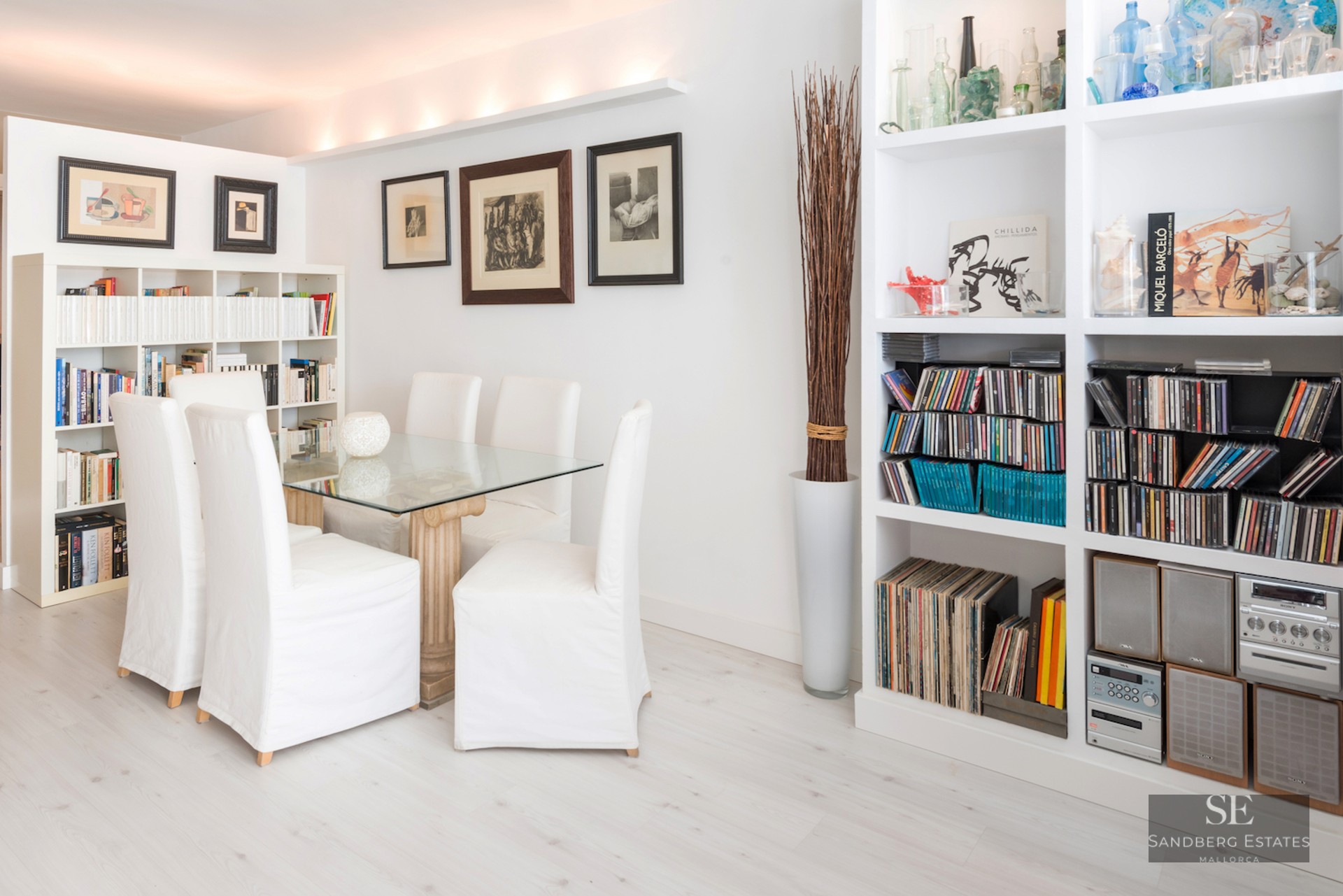A bright dining room with a glass table, white chairs, and extensive white bookshelves filled with books and music.