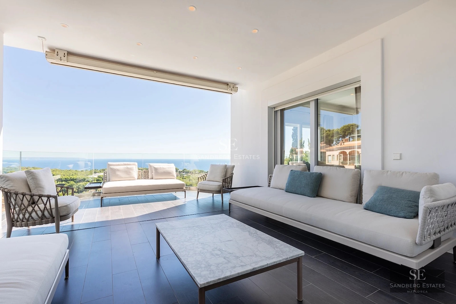 Spacious modern terrace with white sofas, marble table, and glass railing overlooking the sea and forest.