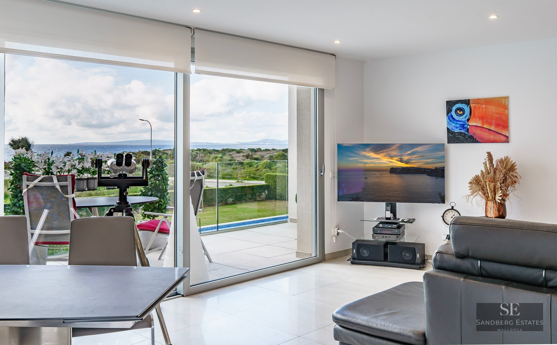 Bright living room with large glass doors opening to a terrace with sea views, featuring a TV and leather sofa.