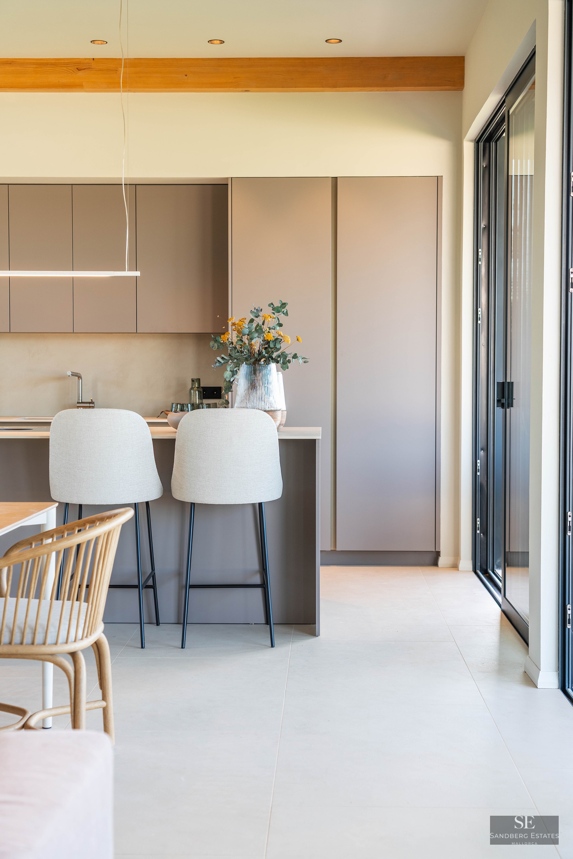 A modern kitchen featuring matte taupe cabinets, an island with two light-colored bar stools, and a wooden ceiling beam.