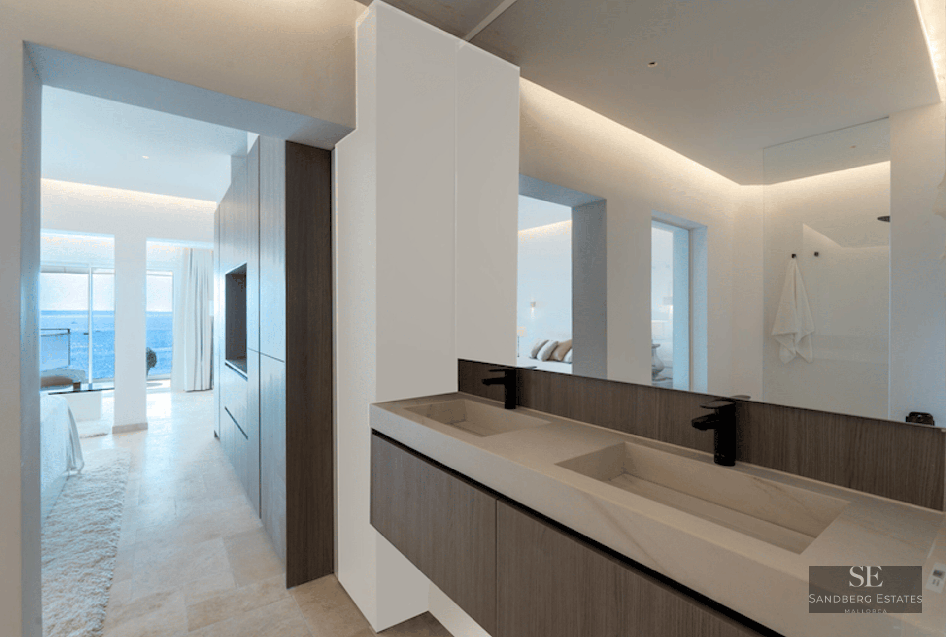 Modern master bathroom featuring double stone sinks, dark wood vanity, and a view through to a bedroom and the sea.