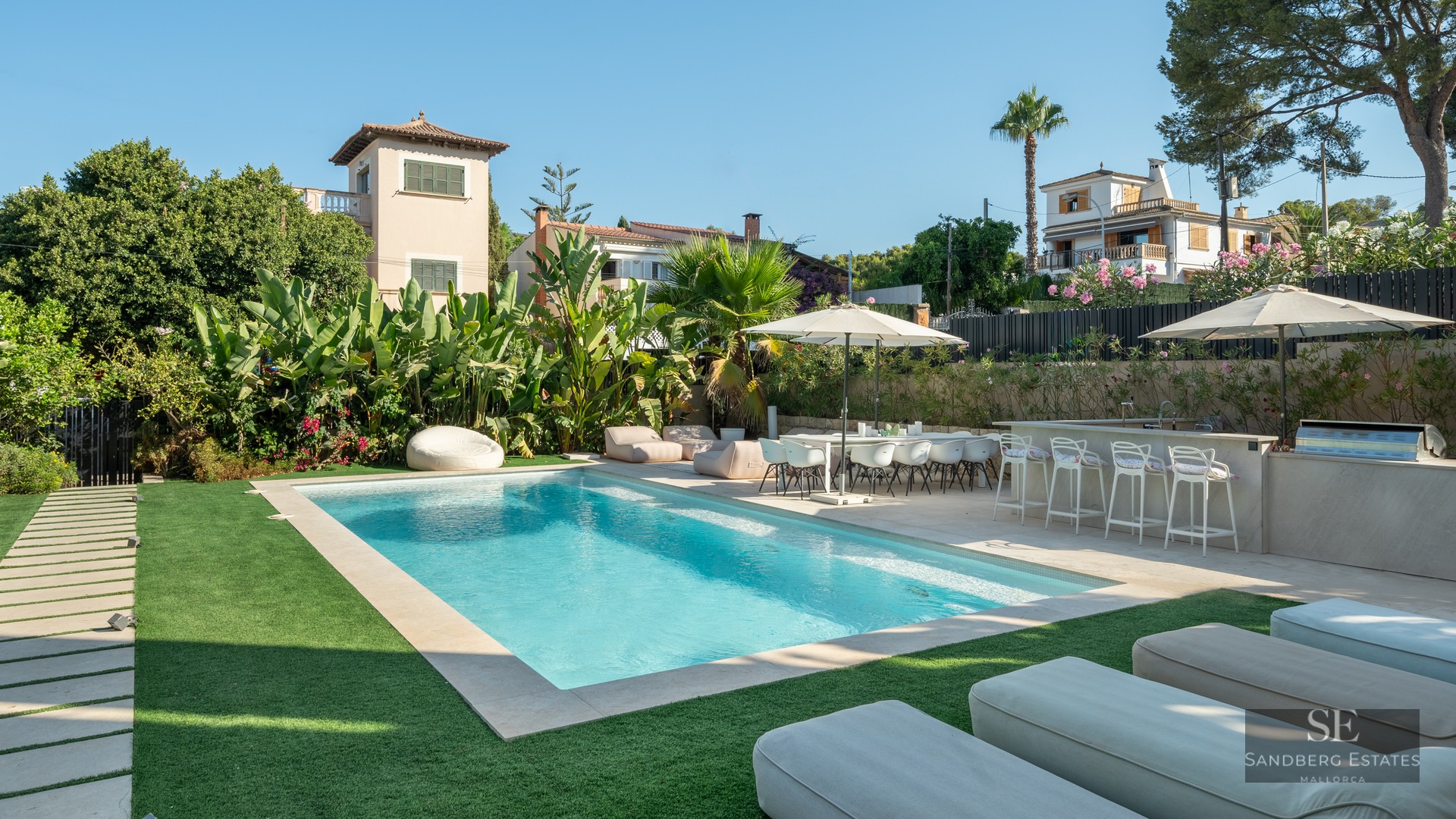 A sparkling rectangular swimming pool surrounded by a green lawn, outdoor dining set, bar, and lush tropical plants.