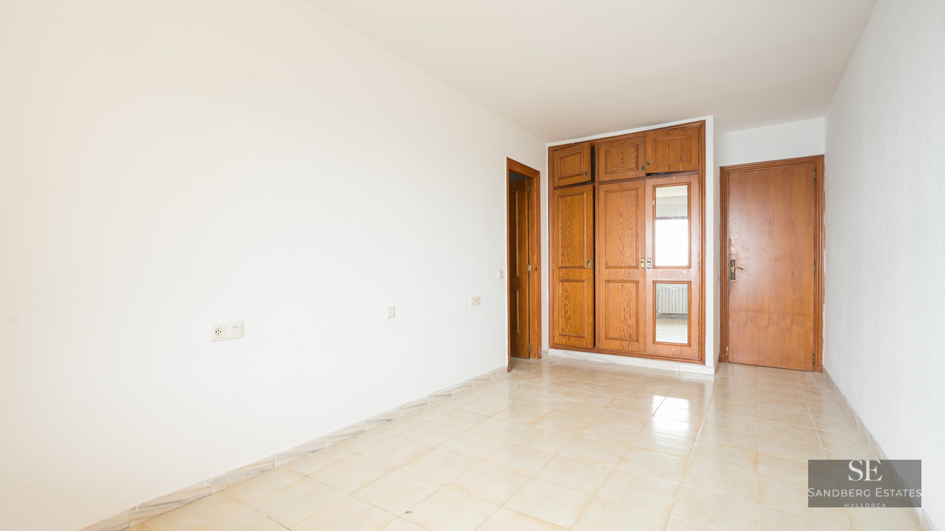 Empty room with white walls, beige tiled floor, and a large built-in wooden wardrobe with a mirrored panel.
