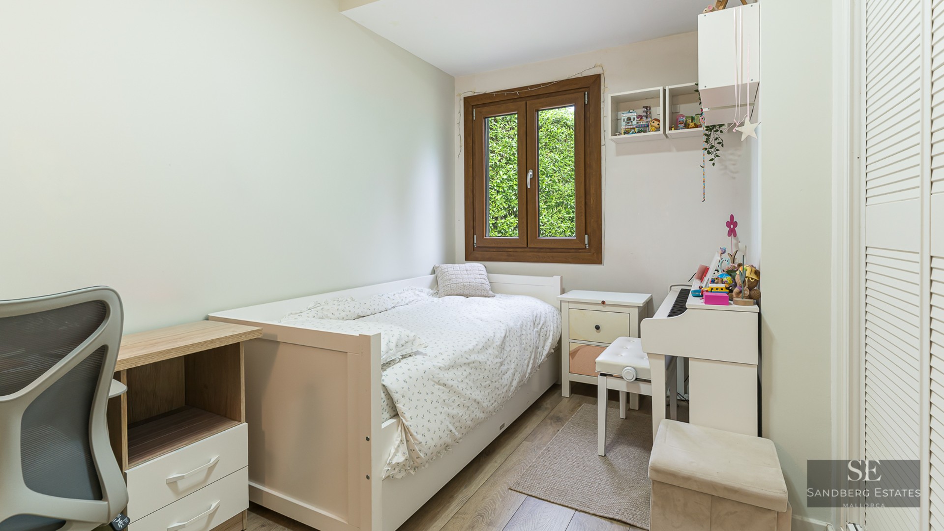 Bright bedroom with a single white bed, a desk, a white digital piano, and a window showing green foliage.