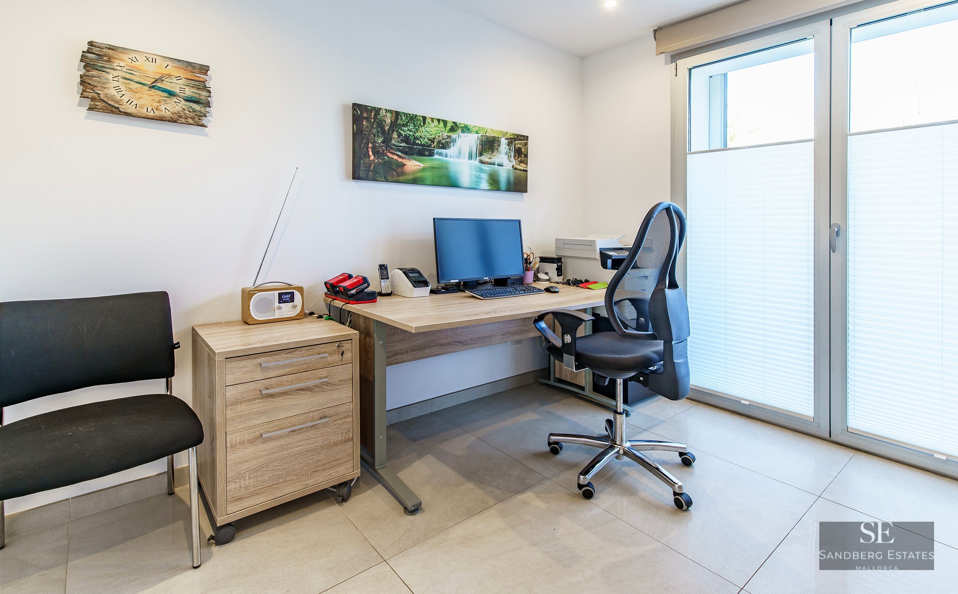 Home office featuring a light wood desk, ergonomic chair, computer setup, and large window with blinds.