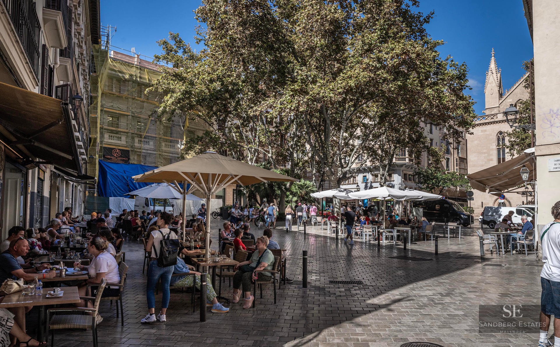 Des gens assis à des tables de café en plein air sous des parasols sur une place pavée avec des arbres et des bâtiments.