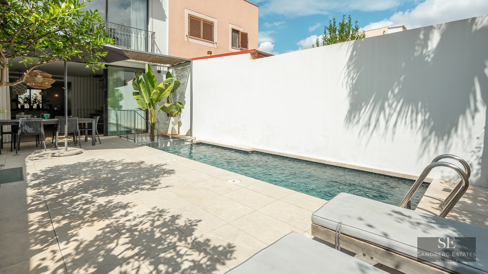 A narrow blue swimming pool on a light-colored tiled terrace with sun loungers and an outdoor dining area.