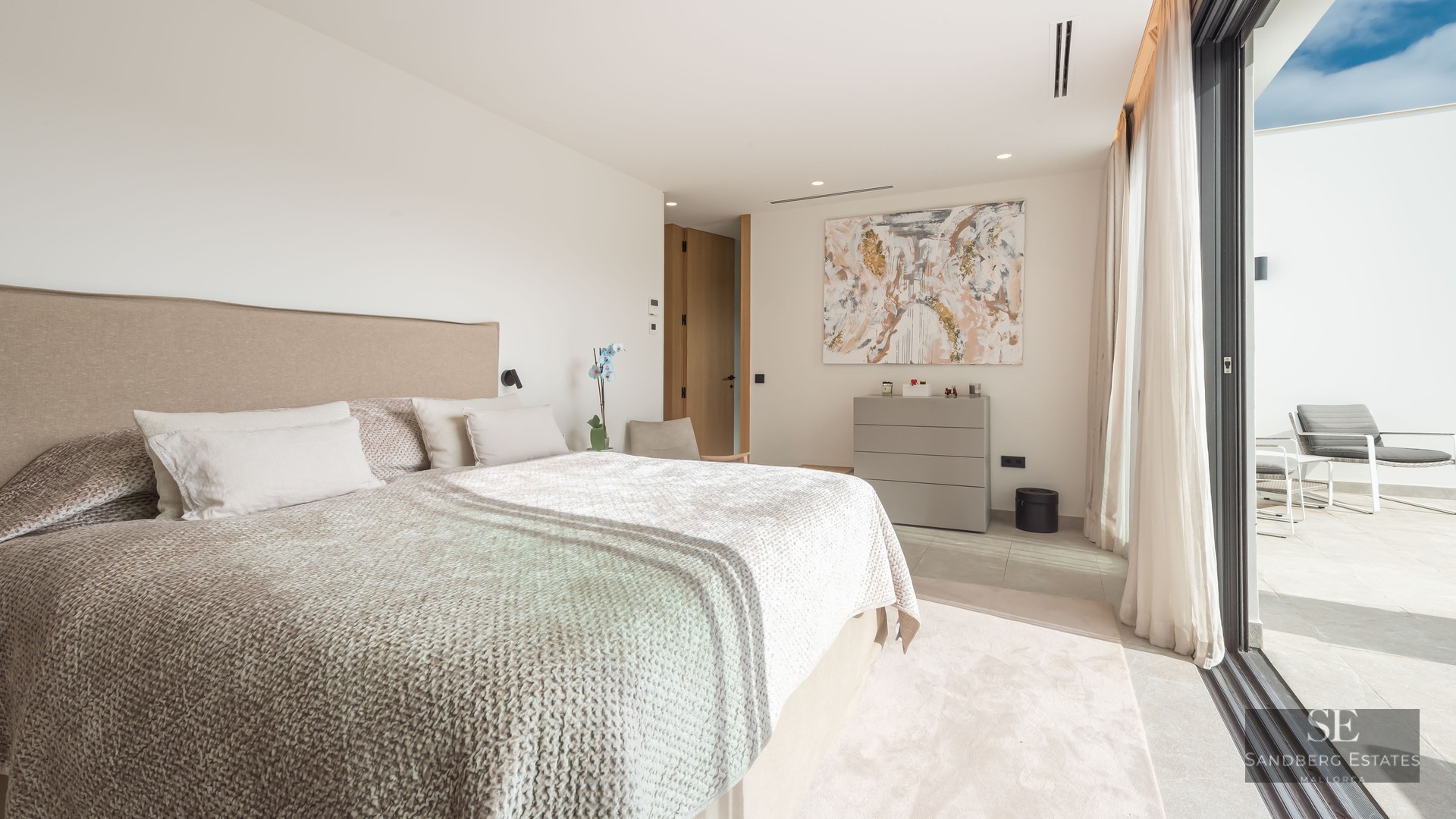 Chambre principale lumineuse avec un grand lit, art abstrait et baies vitrées coulissantes ouvrant sur une terrasse.