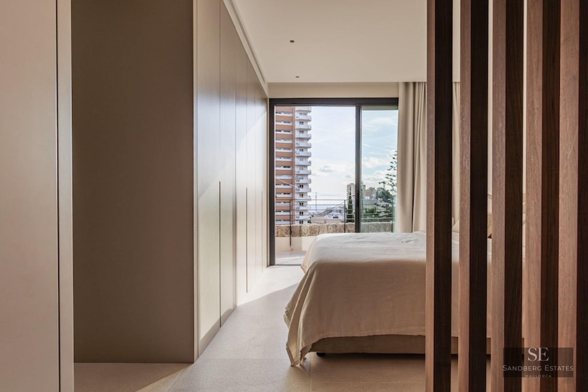 Modern bedroom with large built-in wardrobes, a bed with light linens, and a balcony window showing city buildings.