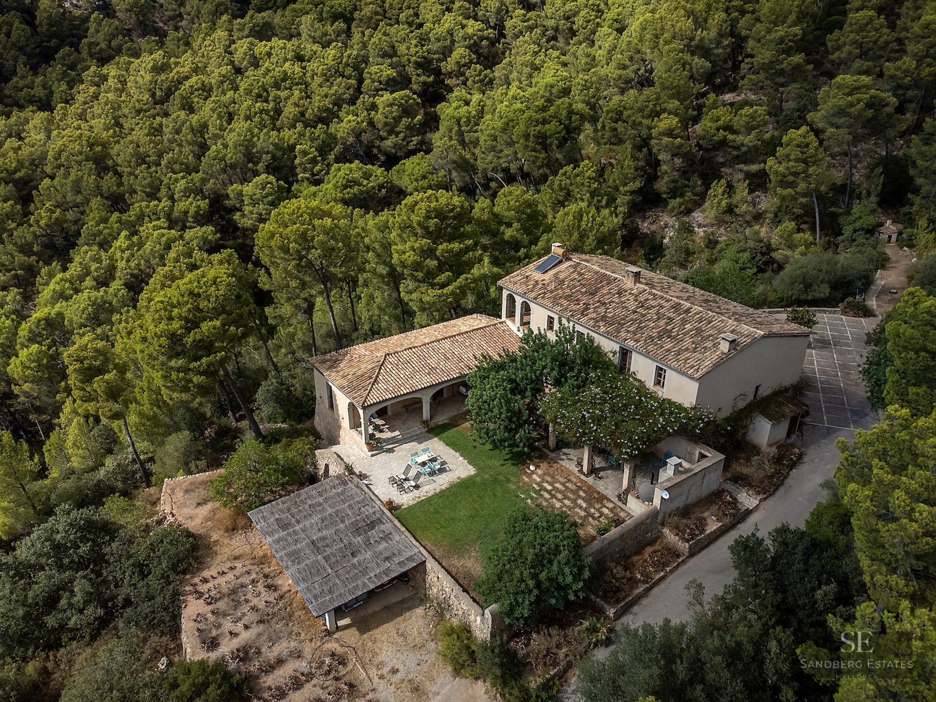 Top-down view of a stone house with tiled roofs, surrounded by a dense green pine forest.