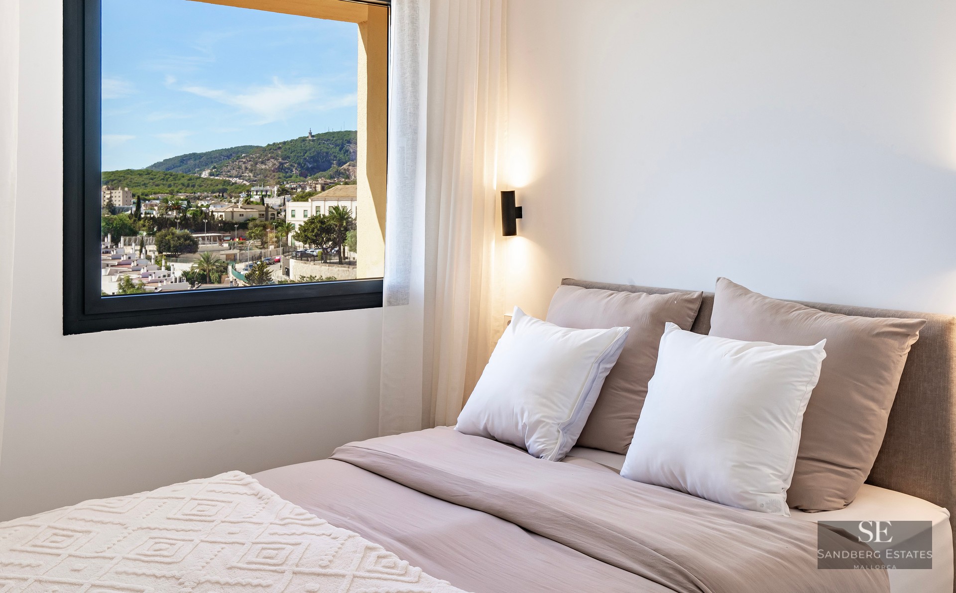 Modern bedroom with neutral-toned bedding and a large window overlooking a coastal town and green hills.