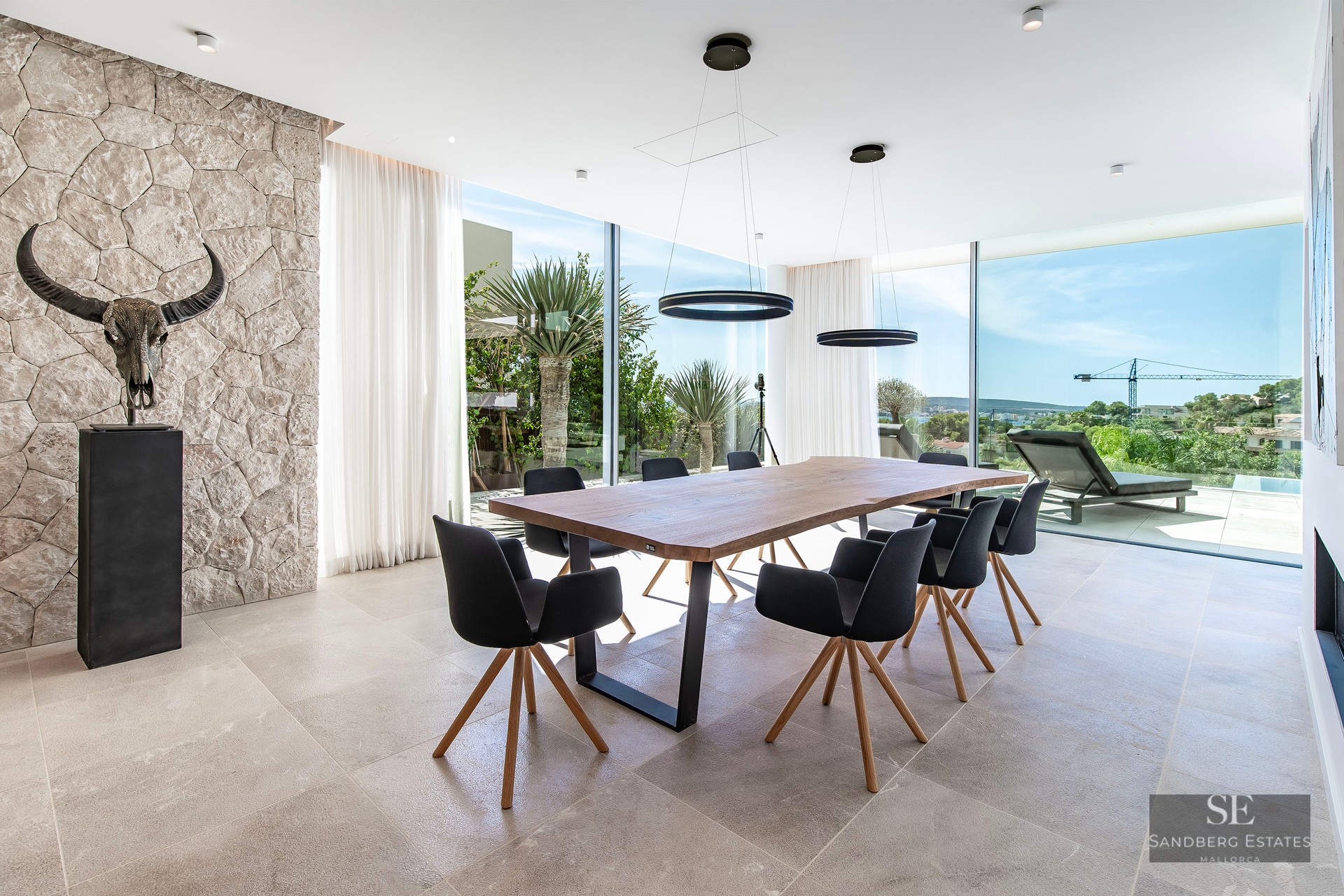 A modern dining room featuring a large wooden table, black chairs, a stone wall with a skull sculpture, and large windows.